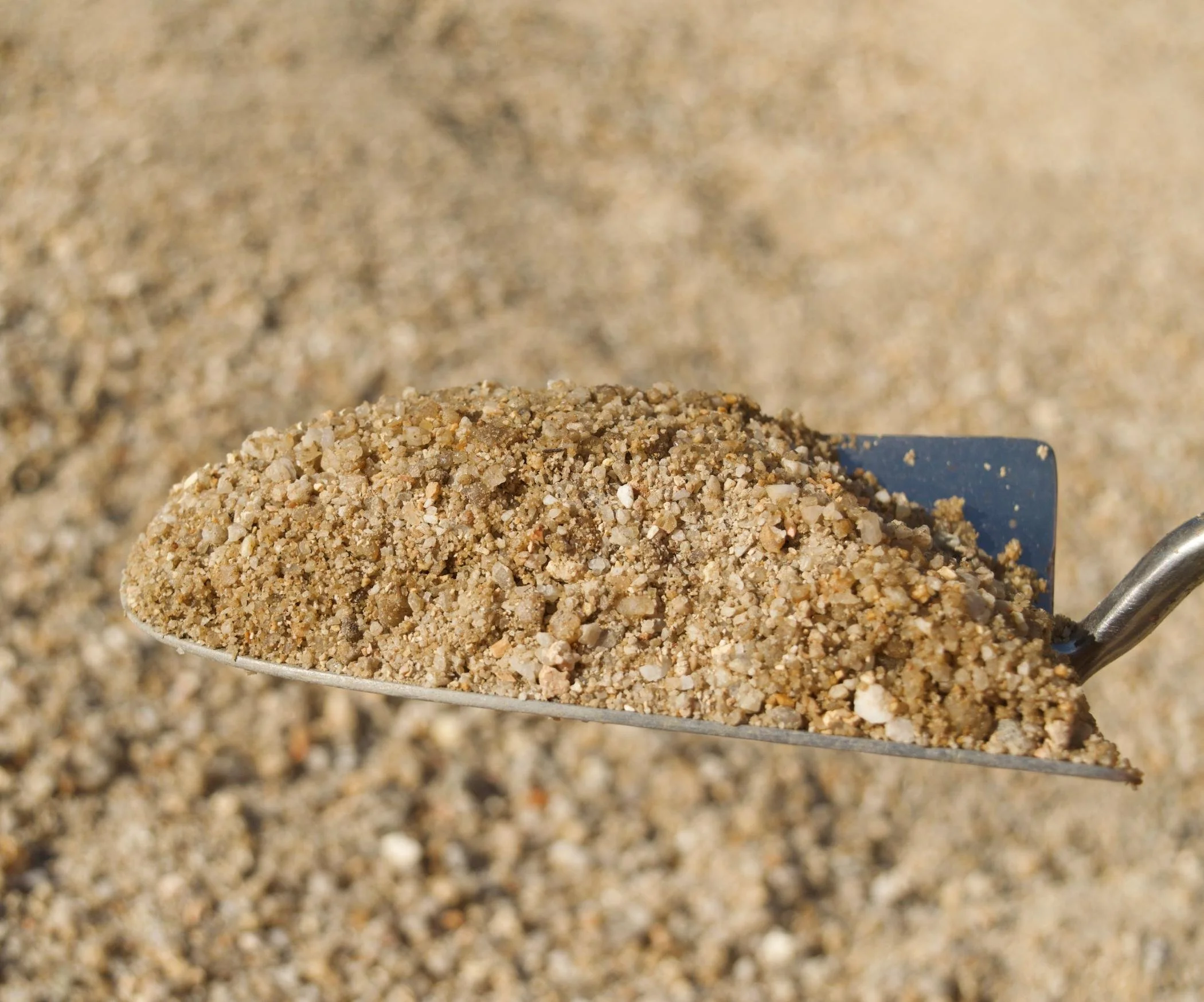 Close-up of a pile of coarse, light-colored sand or gravel on a metallic shovel, with a blurred background of similar material.