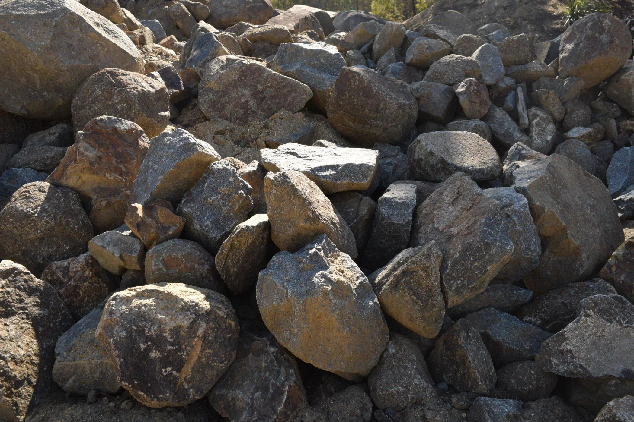 A large pile of rocks and stones outdoors, varying in size, shape, and color with some trees visible in the background.