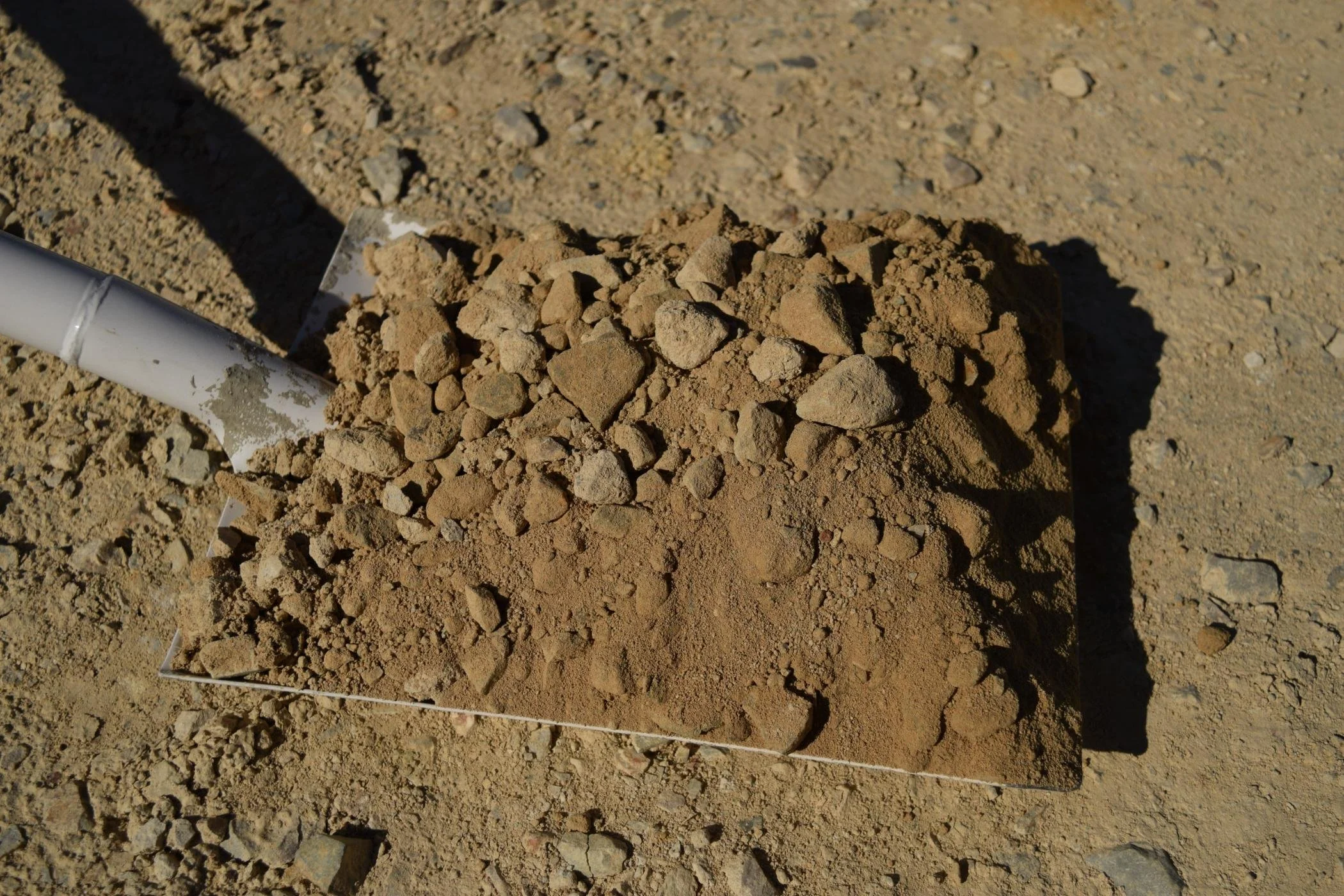 Close-up of a mound of dirt and small rocks on a construction site, with a white plastic pipe and a trowel visible in the scene.