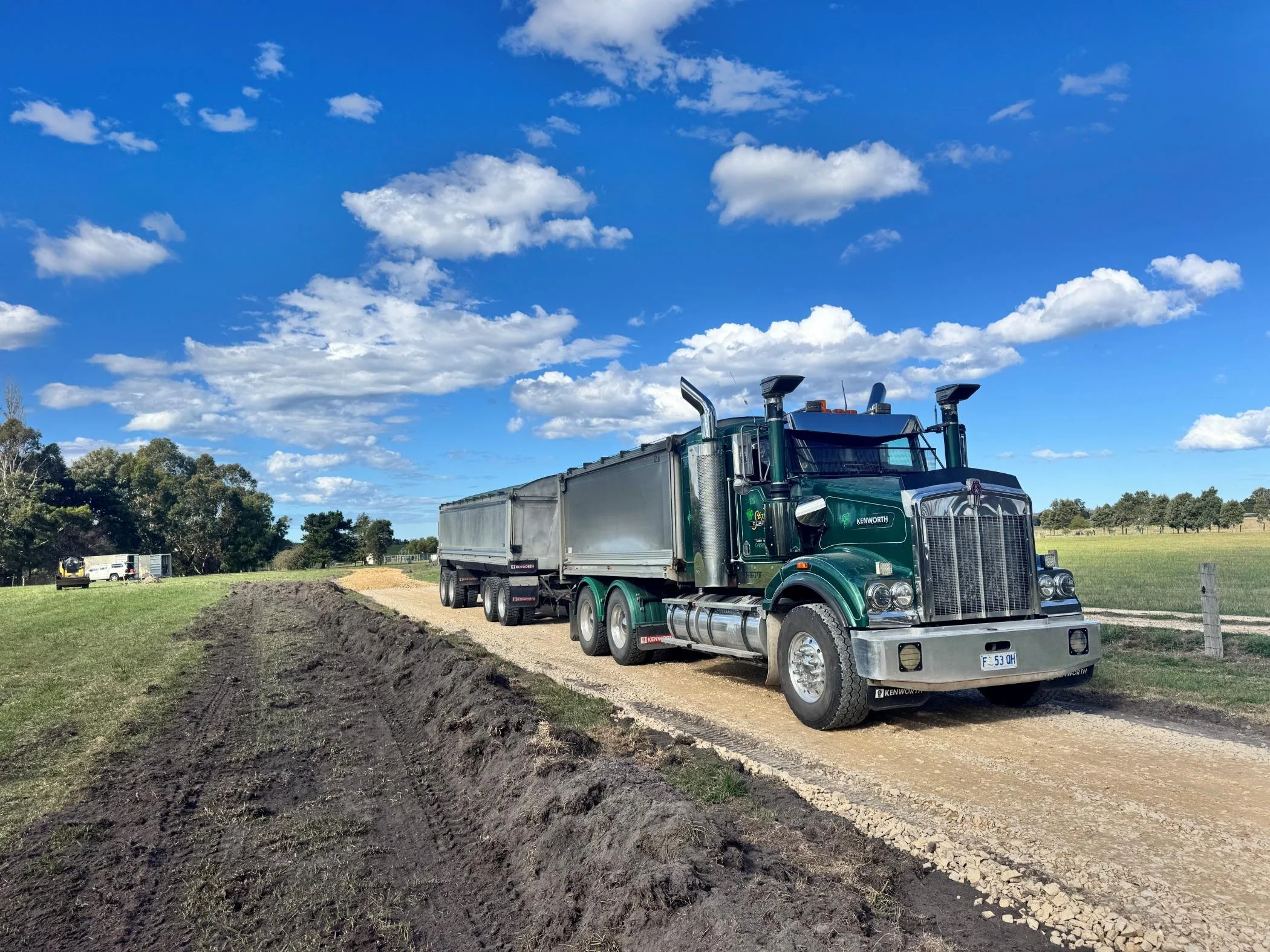 A large green Kenworth semi-truck parked on a dirt road in a rural area with green grass, trees, and a clear blue sky with scattered clouds.