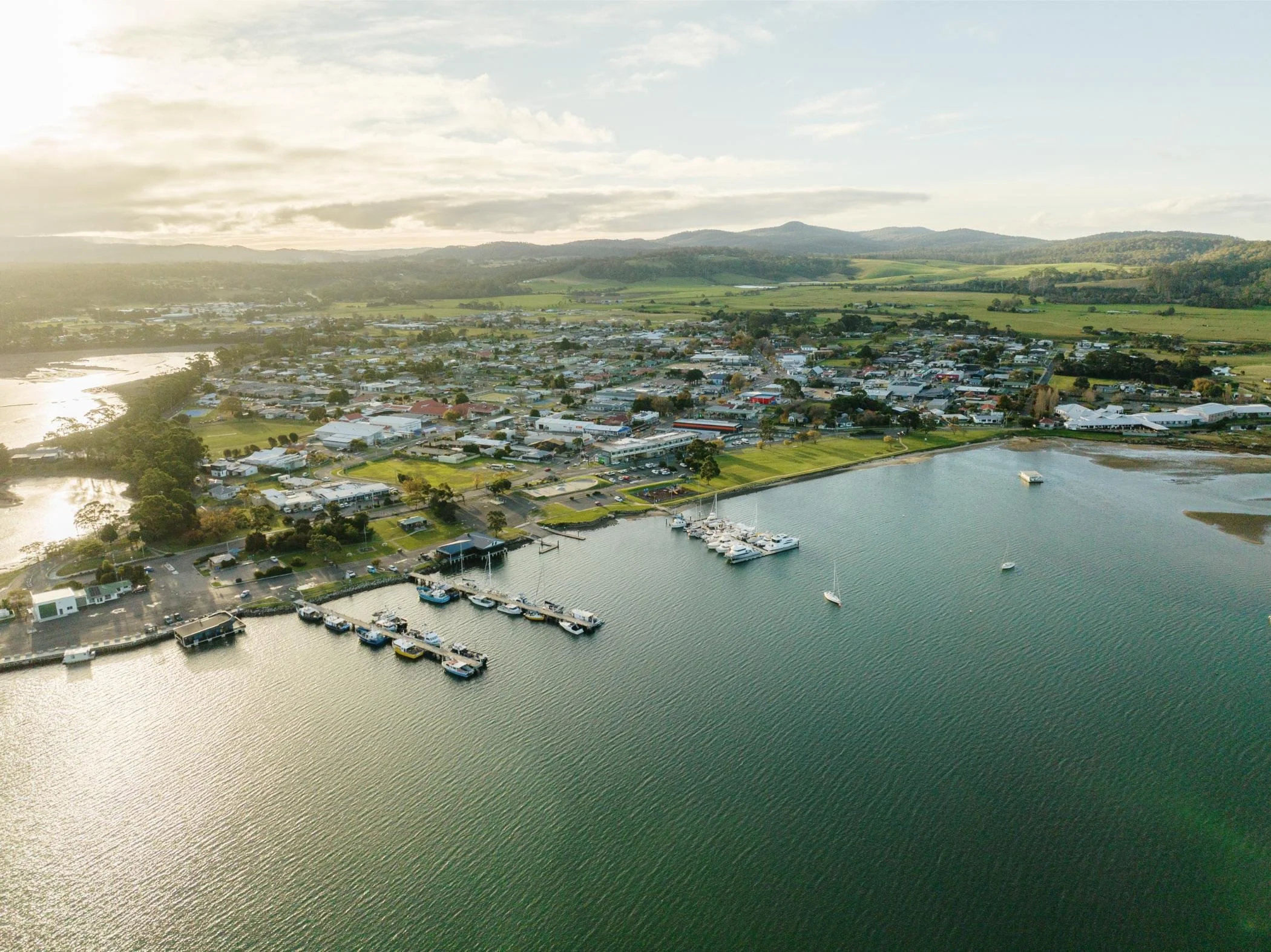 An aerial view of a small town with a marina and boats by a body of water, surrounded by green fields and rolling hills under a partly cloudy sky.