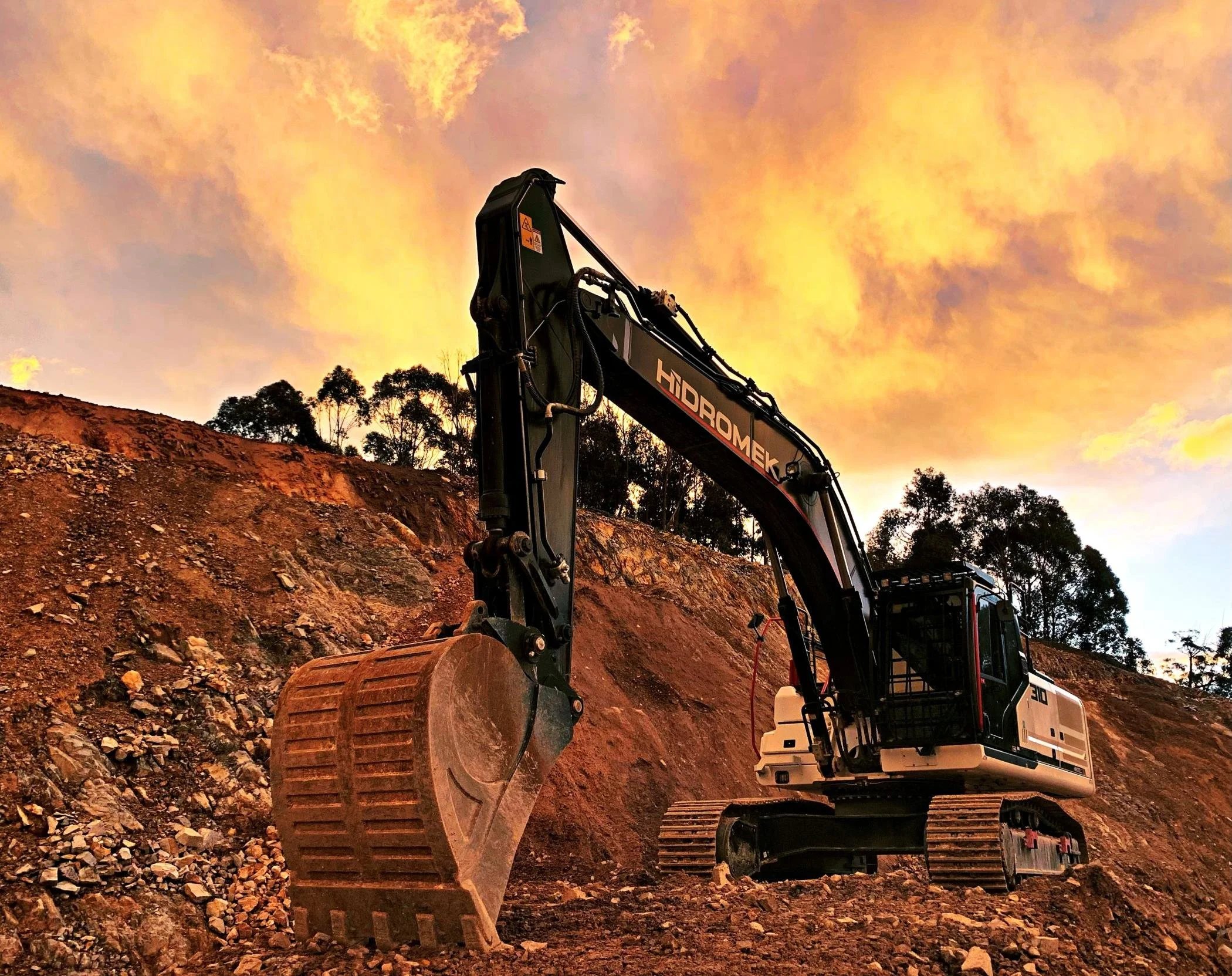 An excavator working on a slope during sunset with a colorful sky and trees in the background.