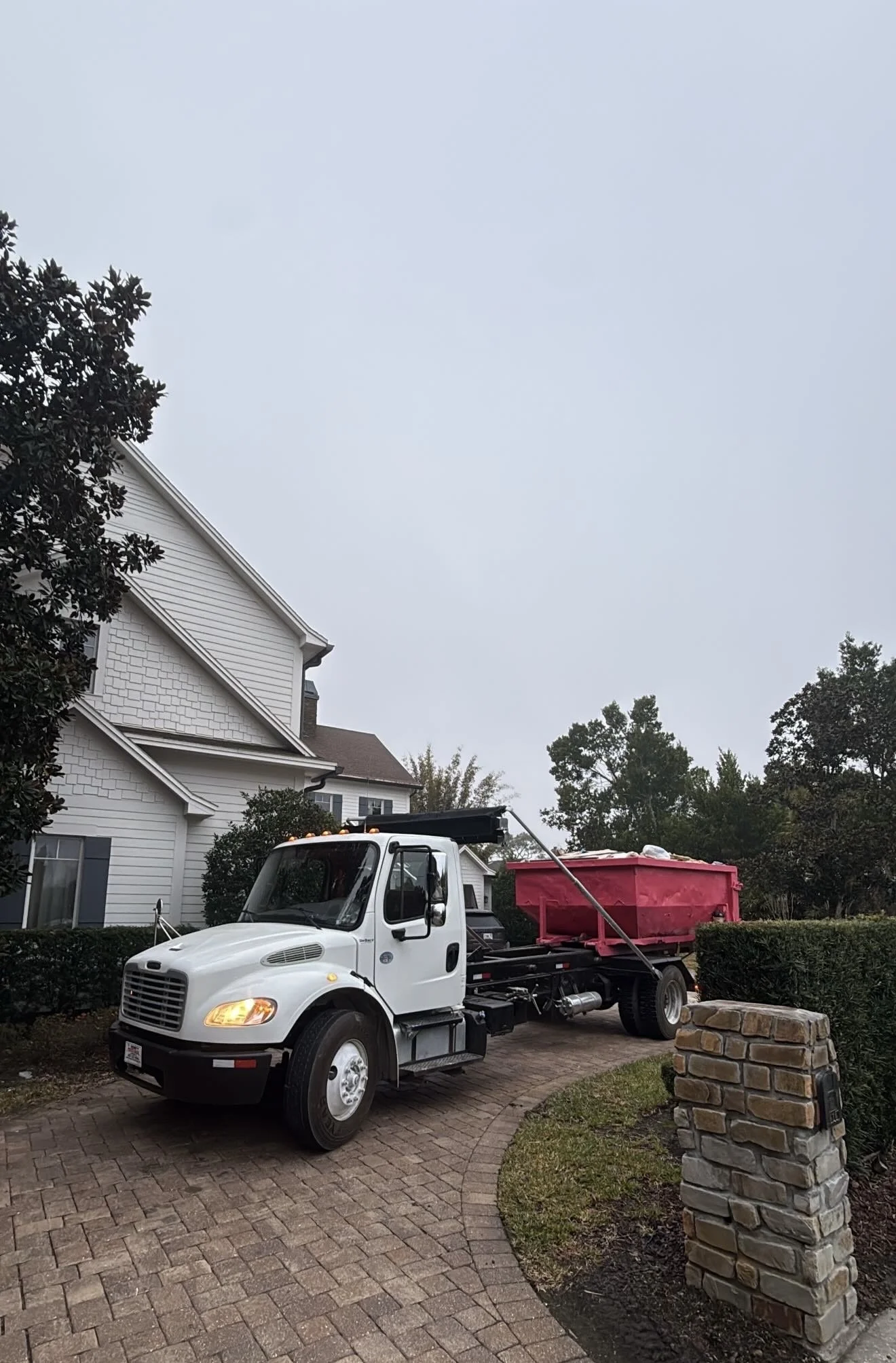 A white and black garbage truck parked on a brick driveway next to a suburban house, with trees and bushes nearby under an overcast sky.
