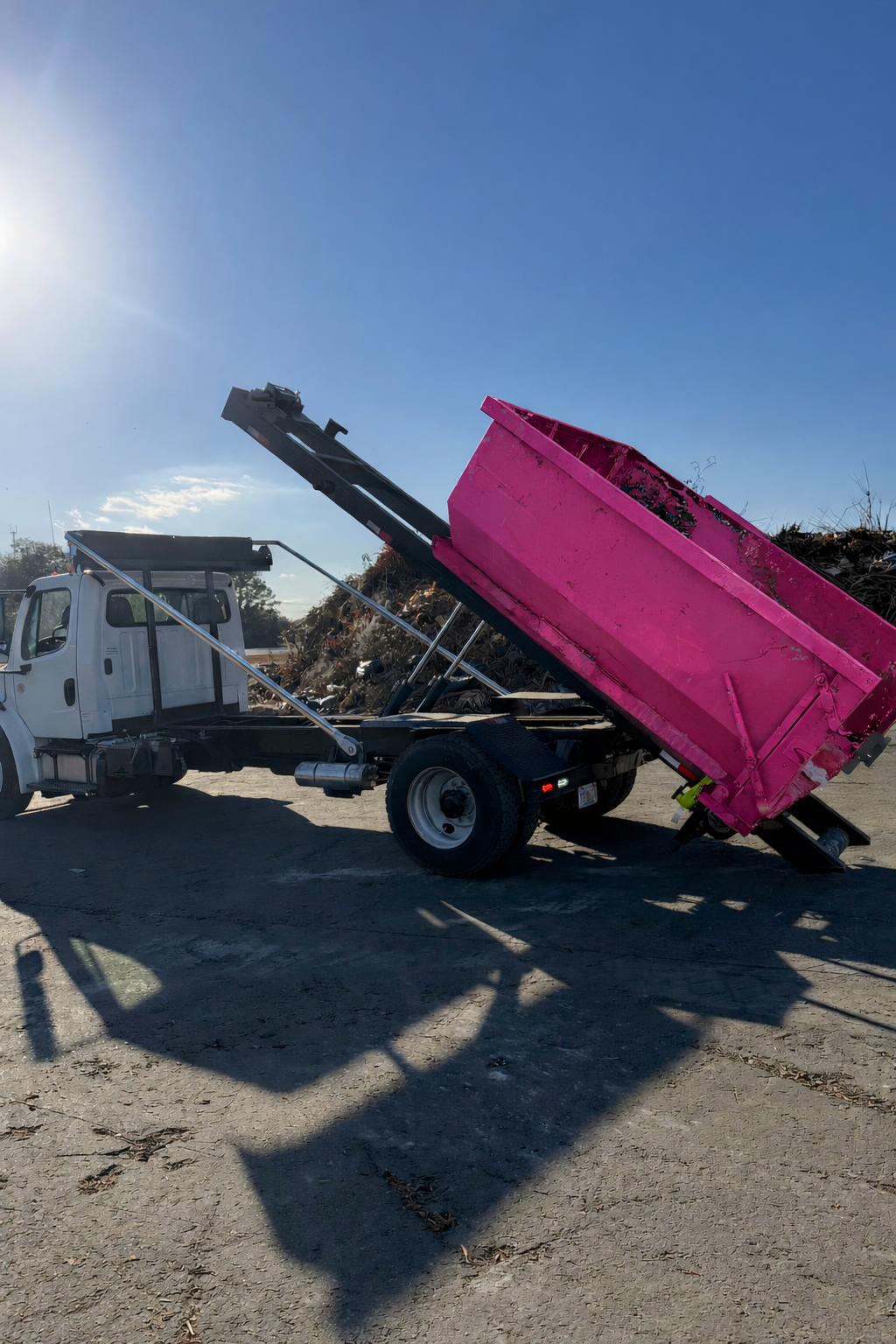 A white dump truck with a pink bed loading debris onto a pile on a gravel lot, under a sunny blue sky.