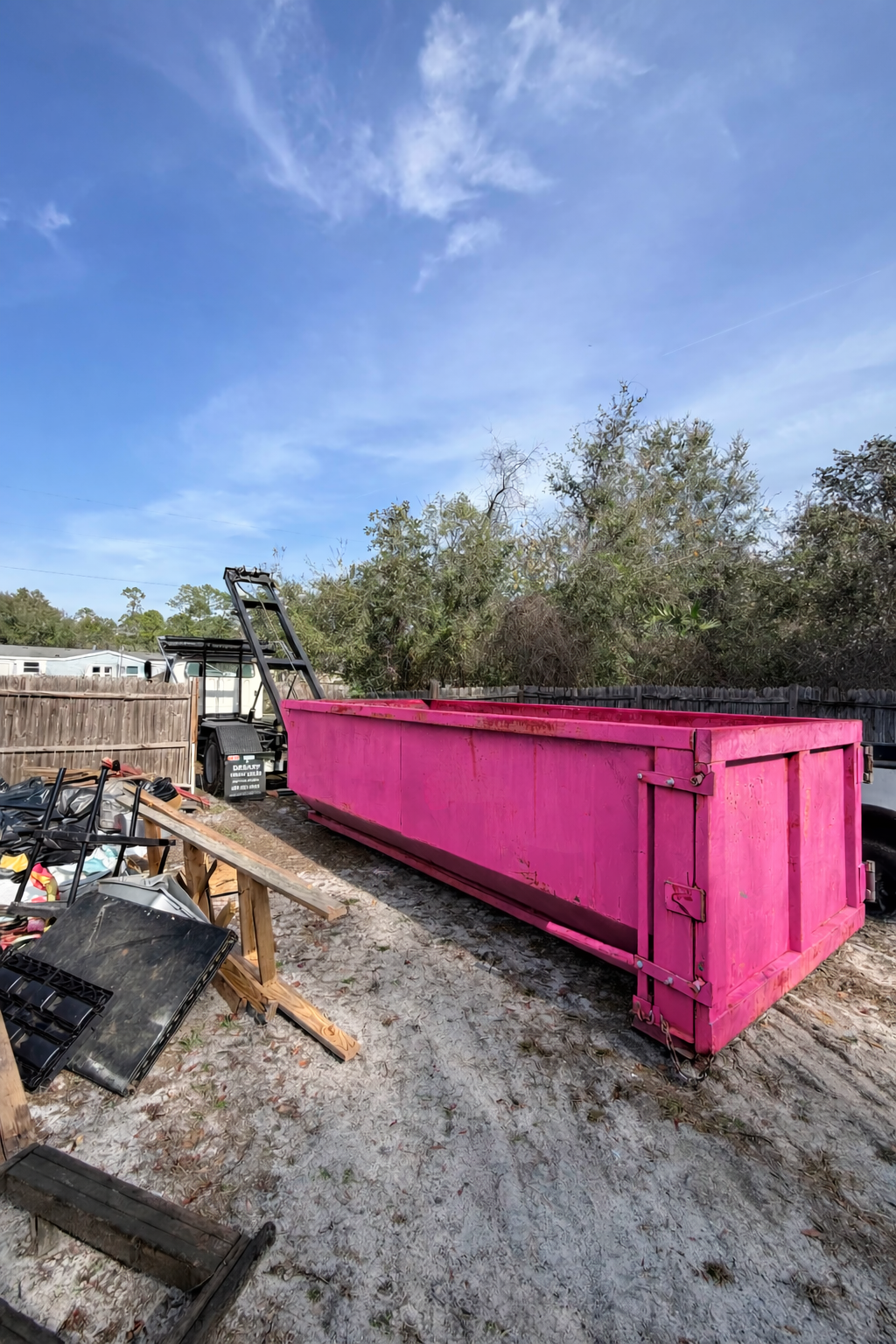 A large pink dumpster in an outdoor yard area with discarded items, a wooden fence, and trees in the background.