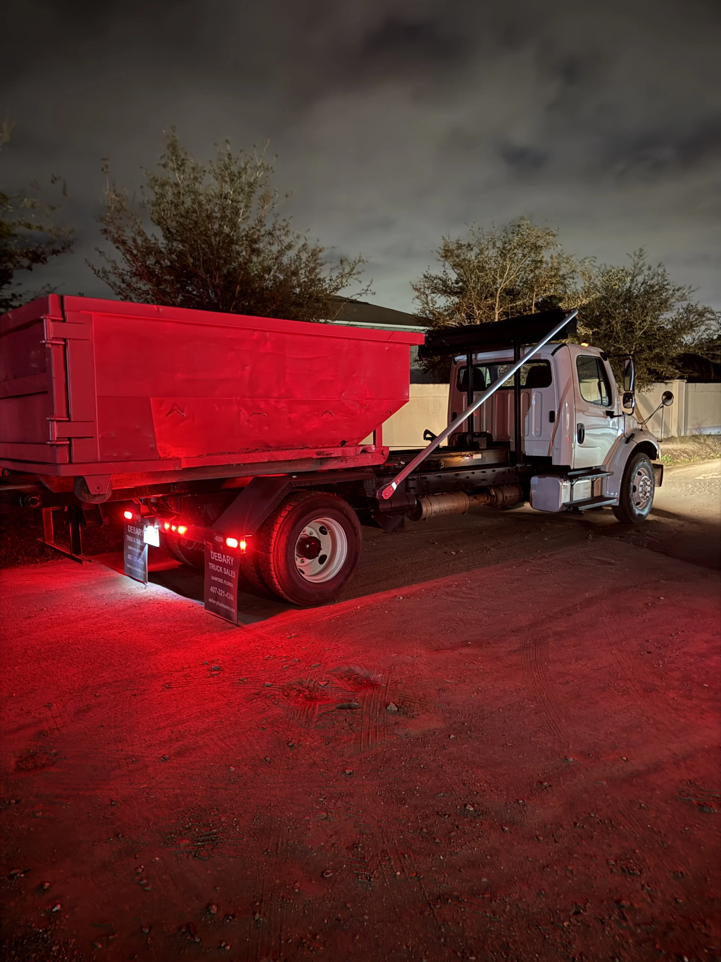 A white flatbed truck with a red cargo container parked on a dirt lot at night, illuminated by red lights with trees and cloudy sky in the background.