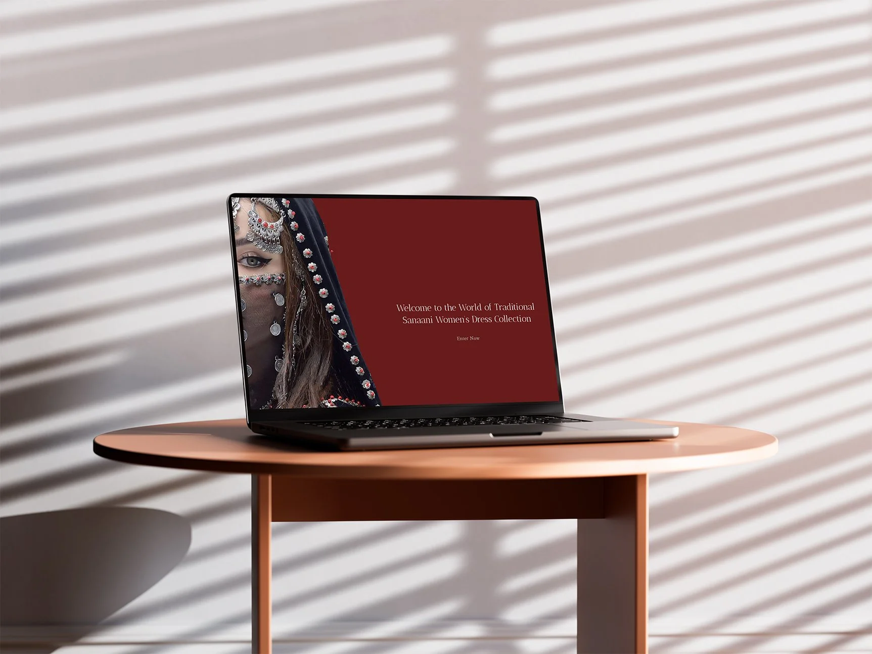 A laptop on a pink round table displaying a welcome message to the World of Traditional Sanaani Women's Dress Collection with a woman in traditional Yemeni attire on the screen, against a background of striped shadows on the wall.