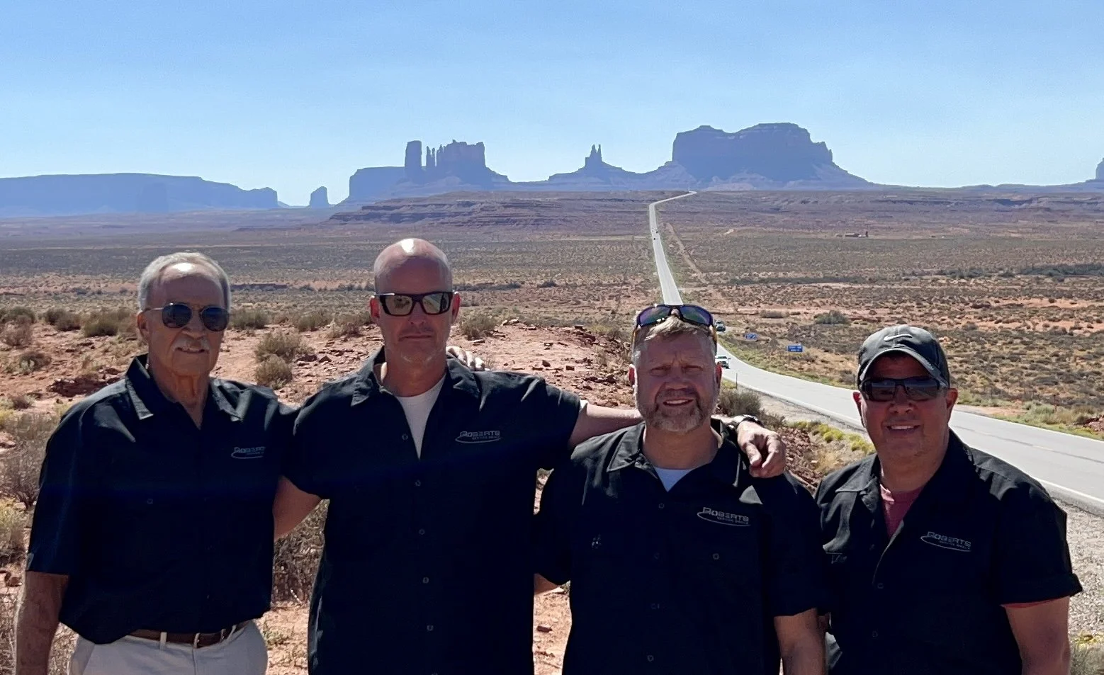 Four men stand together in a desert landscape with a road and Monument Valley in the background.