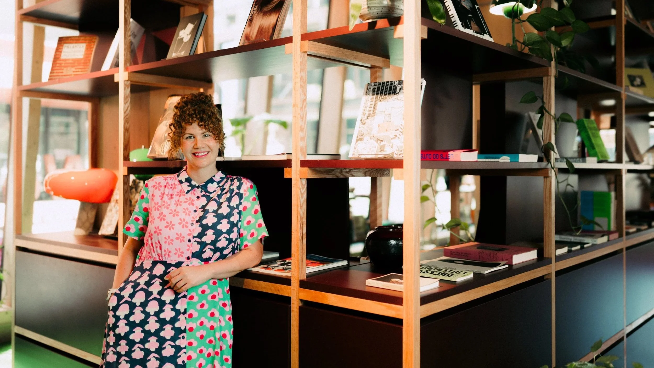 Smiling woman in a vibrant patterned dress leaning against a wooden bookshelf filled with books and greenery in a warm, welcoming space.