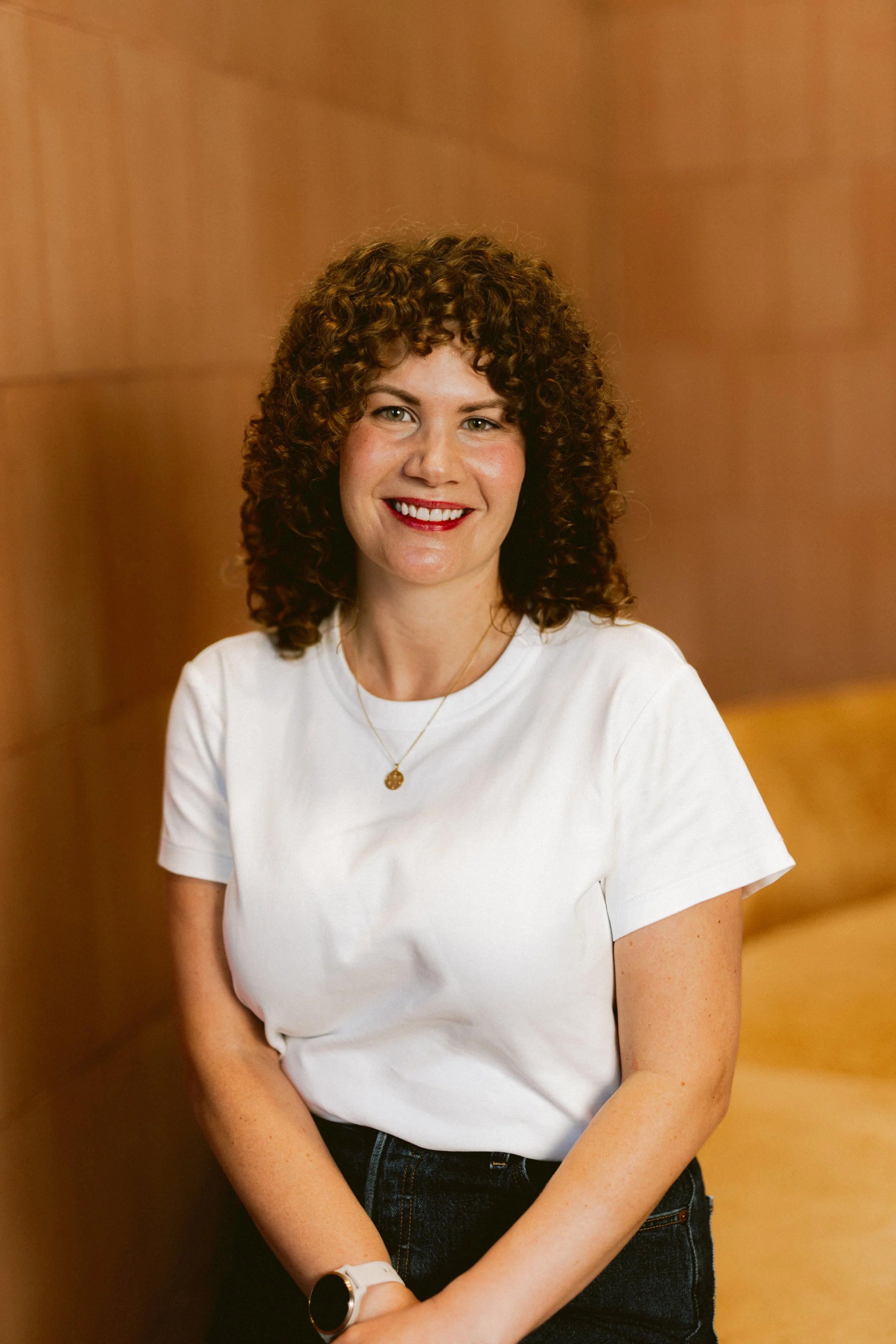 Smiling woman with curly hair in a white T-shirt, standing against a warm timber backdrop in a welcoming space.