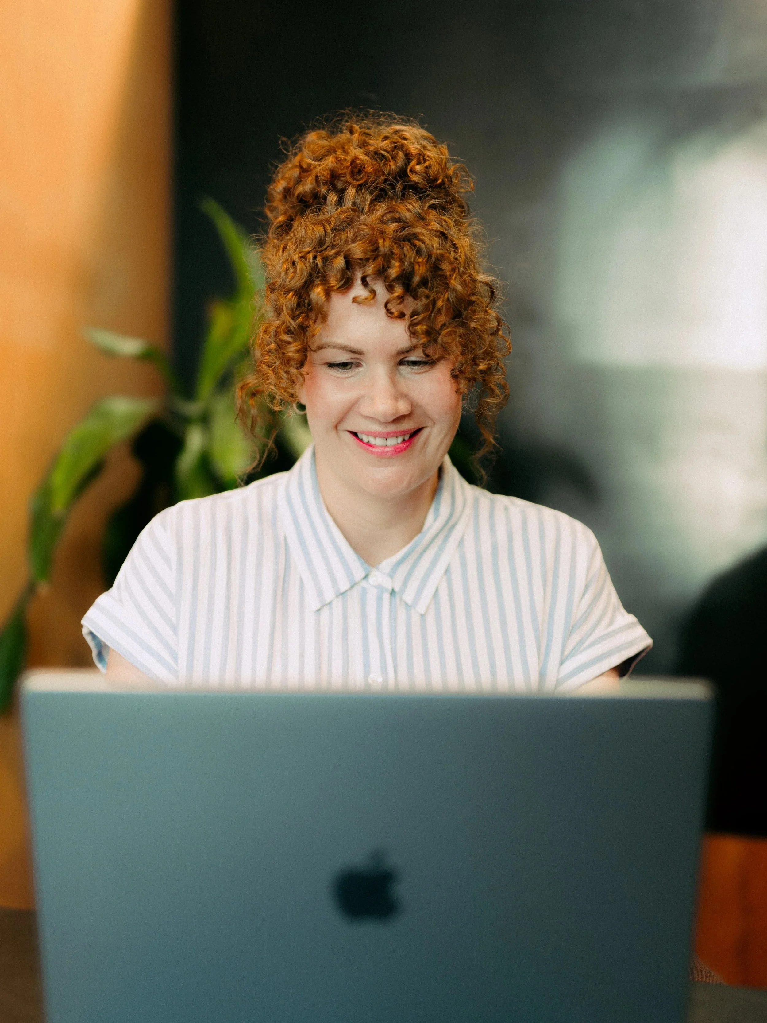 Smiling woman with curly hair working on a laptop in a warm, softly lit space.