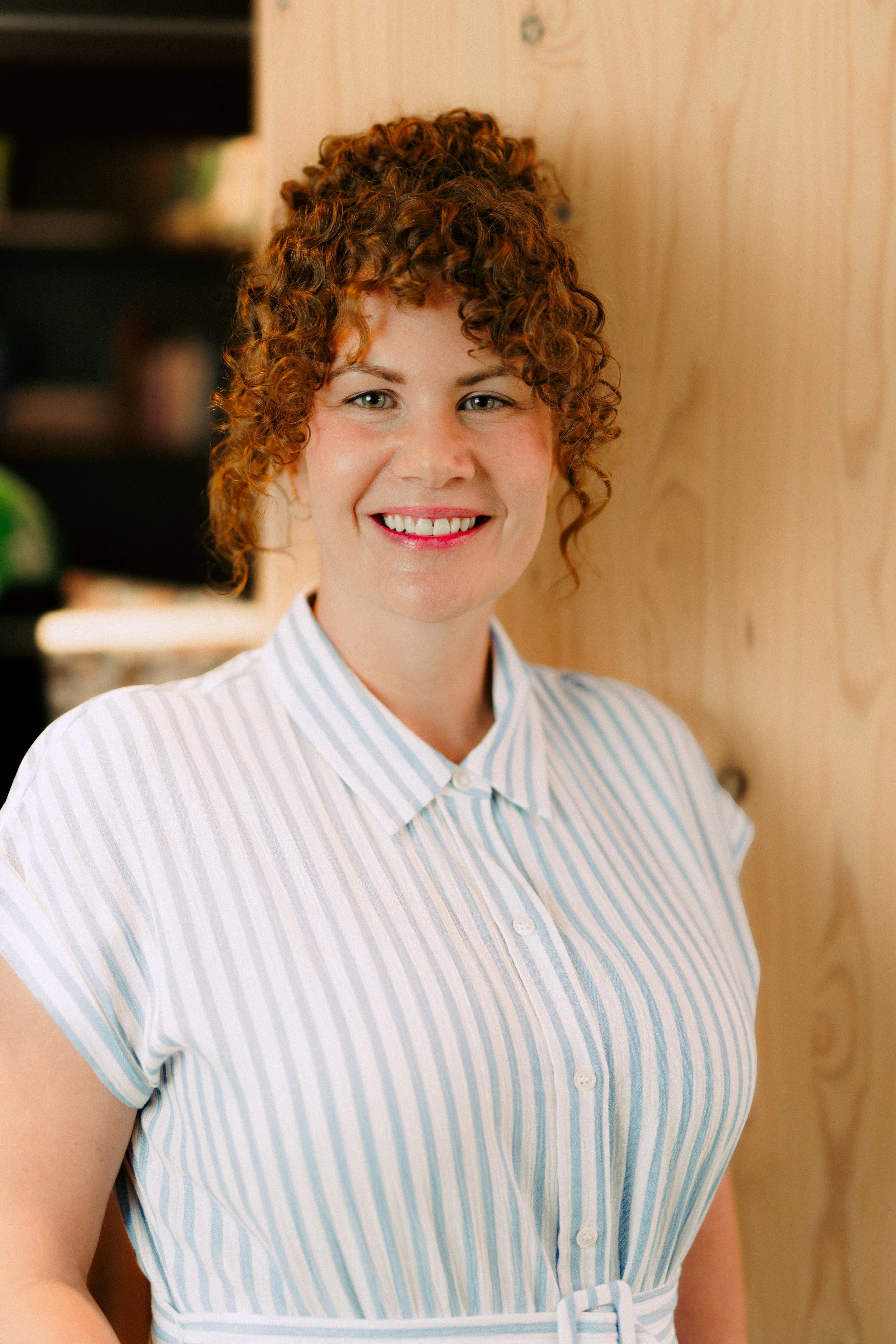 Smiling woman with curly hair in a light blue striped dress, standing against a warm timber backdrop in a bright, welcoming space.