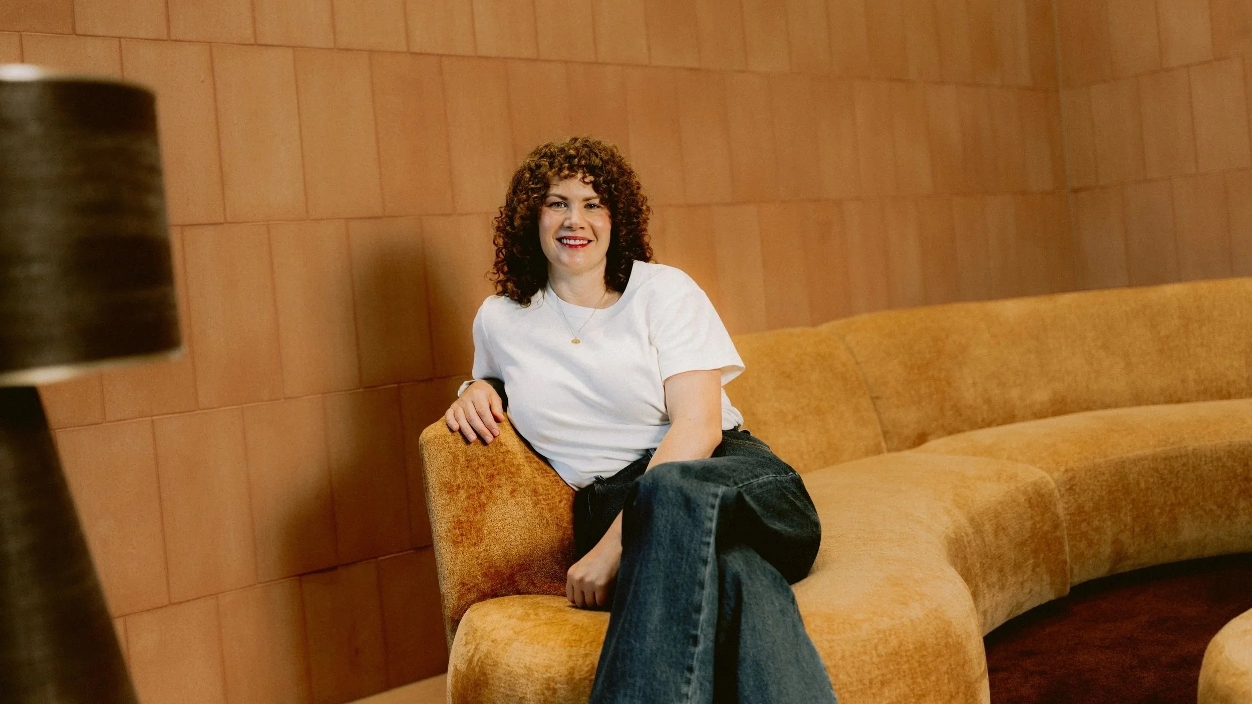 Smiling woman with curly hair seated on a curved mustard sofa in a warm, softly lit space with timber walls.