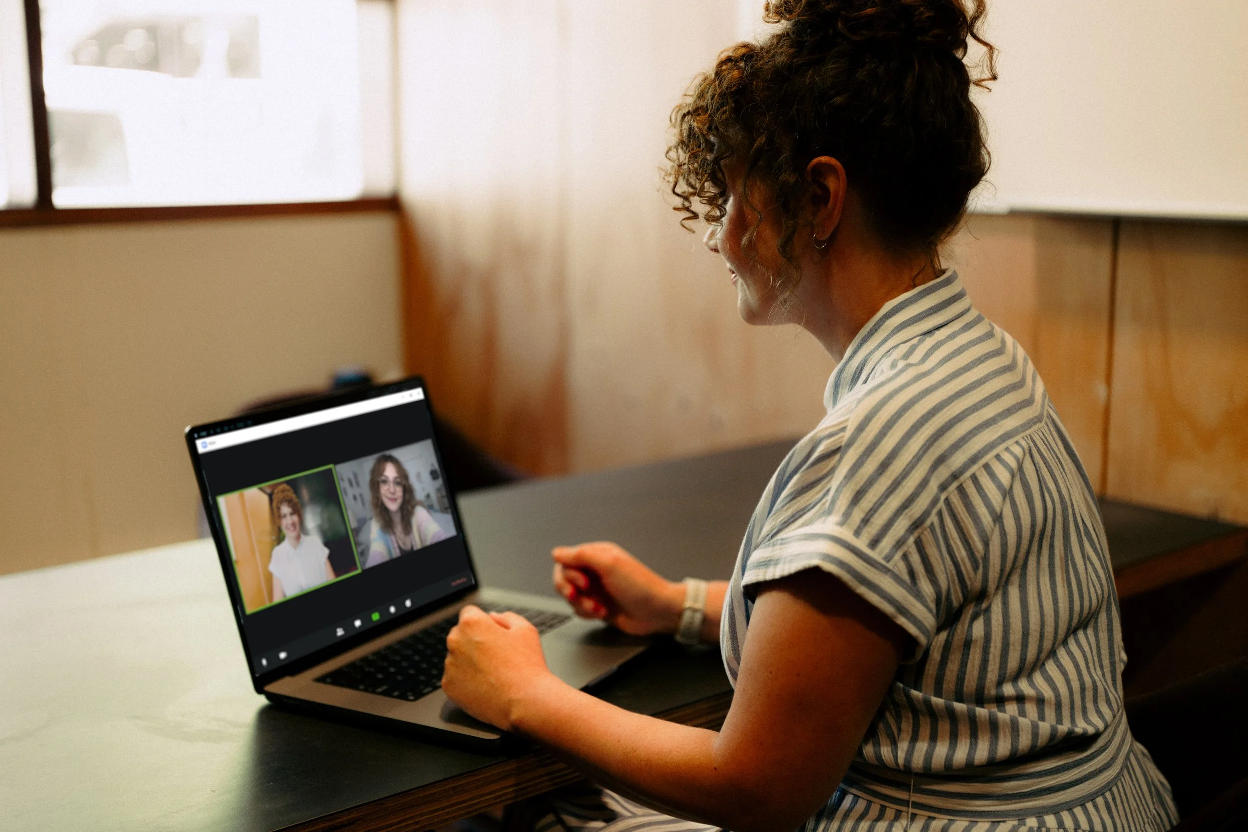 Woman with curly hair sitting at a desk, participating in a video call on her laptop.