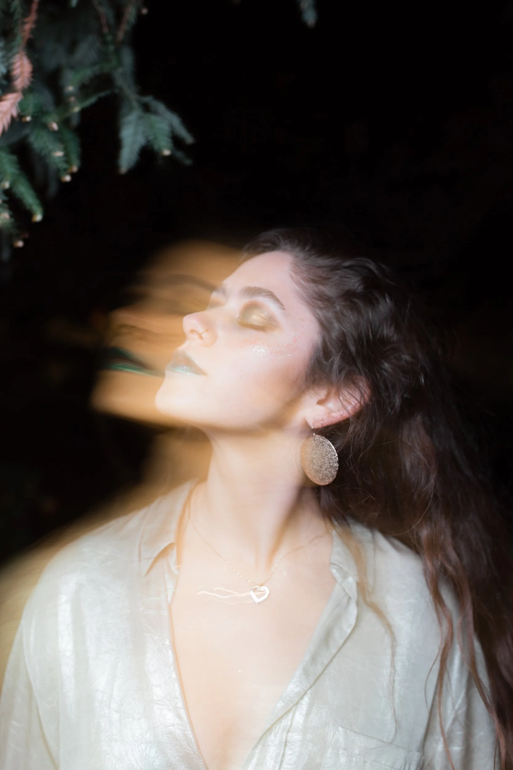 A woman with closed eyes and flowing hair, wearing glittery makeup, large earrings, a heart-shaped necklace, and a shiny top, standing against a black background with a Christmas tree visible in the upper left corner.