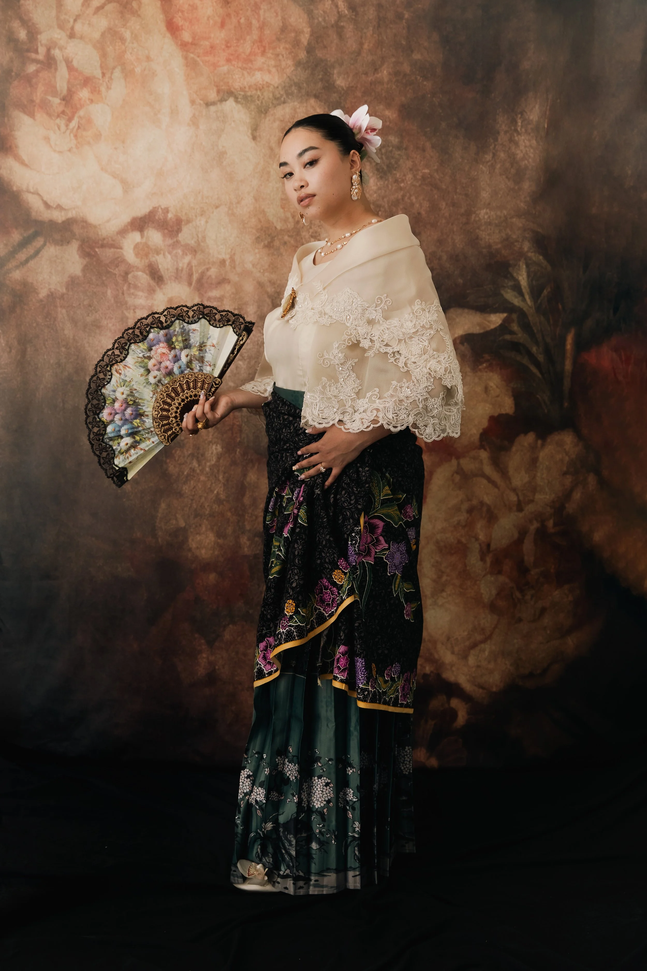 A woman dressed in traditional Indonesian attire, holding a decorative fan, standing against a painted floral background.