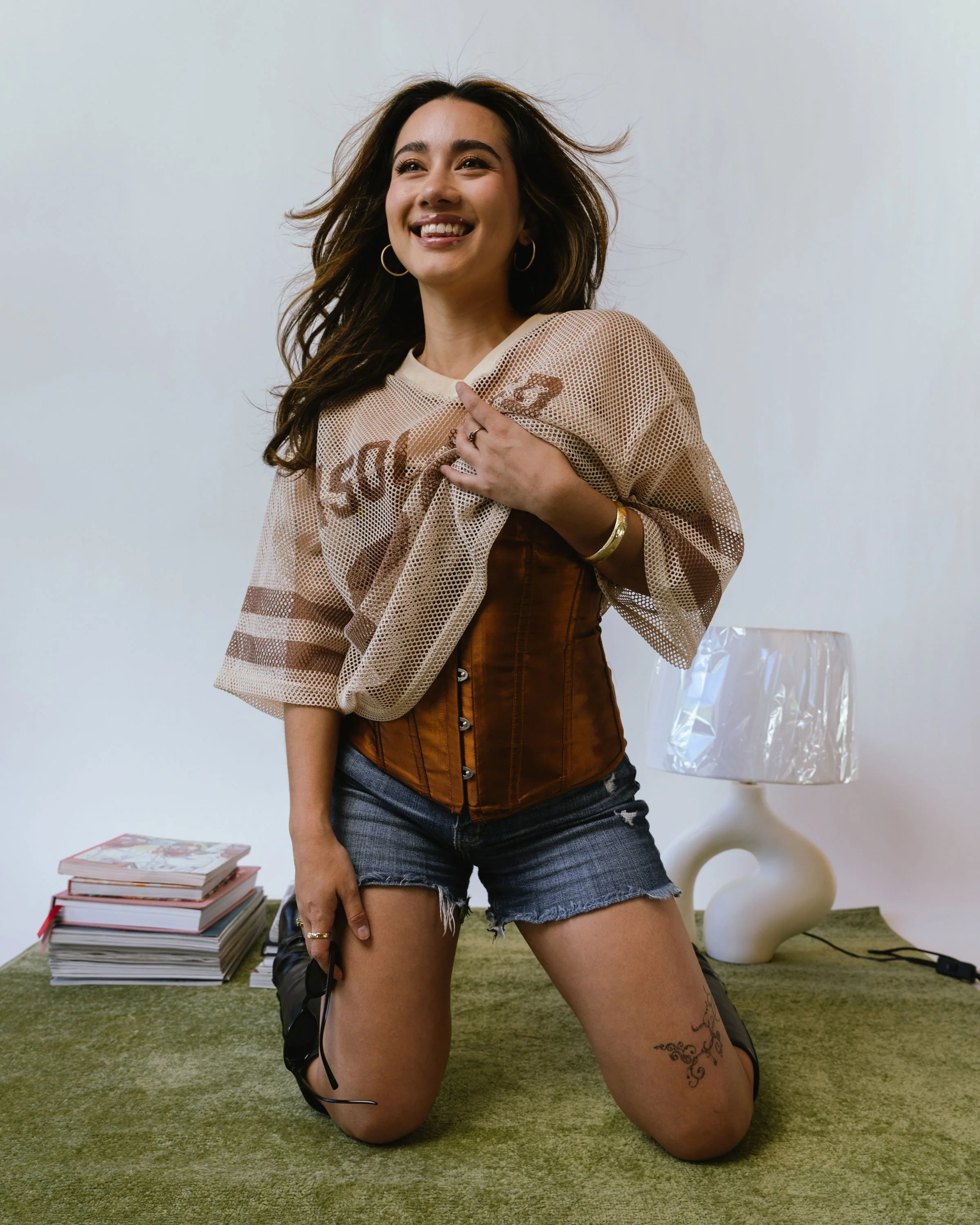 A woman with long dark hair wearing a beige mesh top, a brown corset, and denim shorts, kneeling on a green carpet in a room with a white wall, a white ceramic lamp, and a stack of magazines in the background.