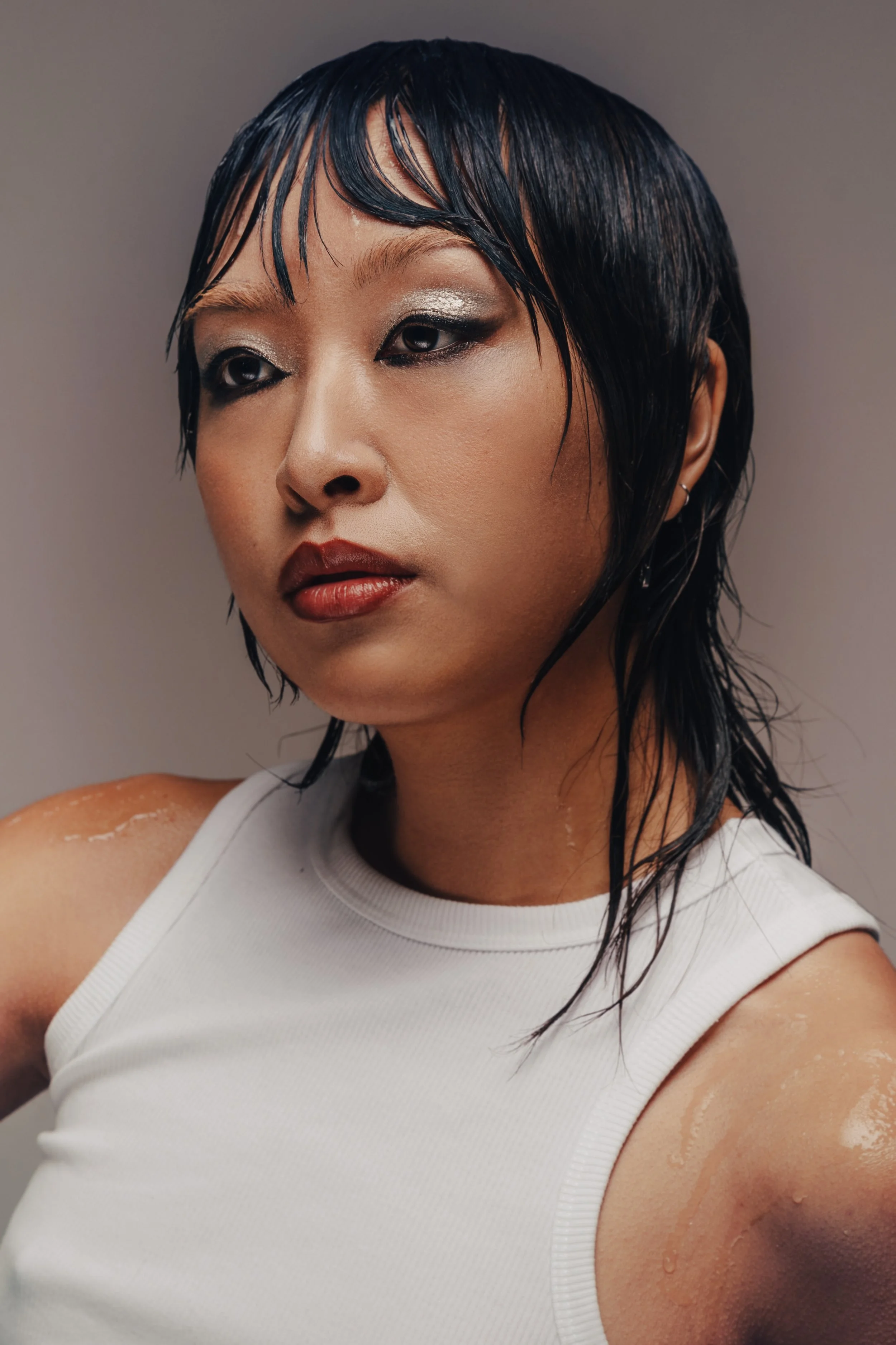 A woman with black, wet hair, wearing a white sleeveless top, looks to the side with a neutral expression.