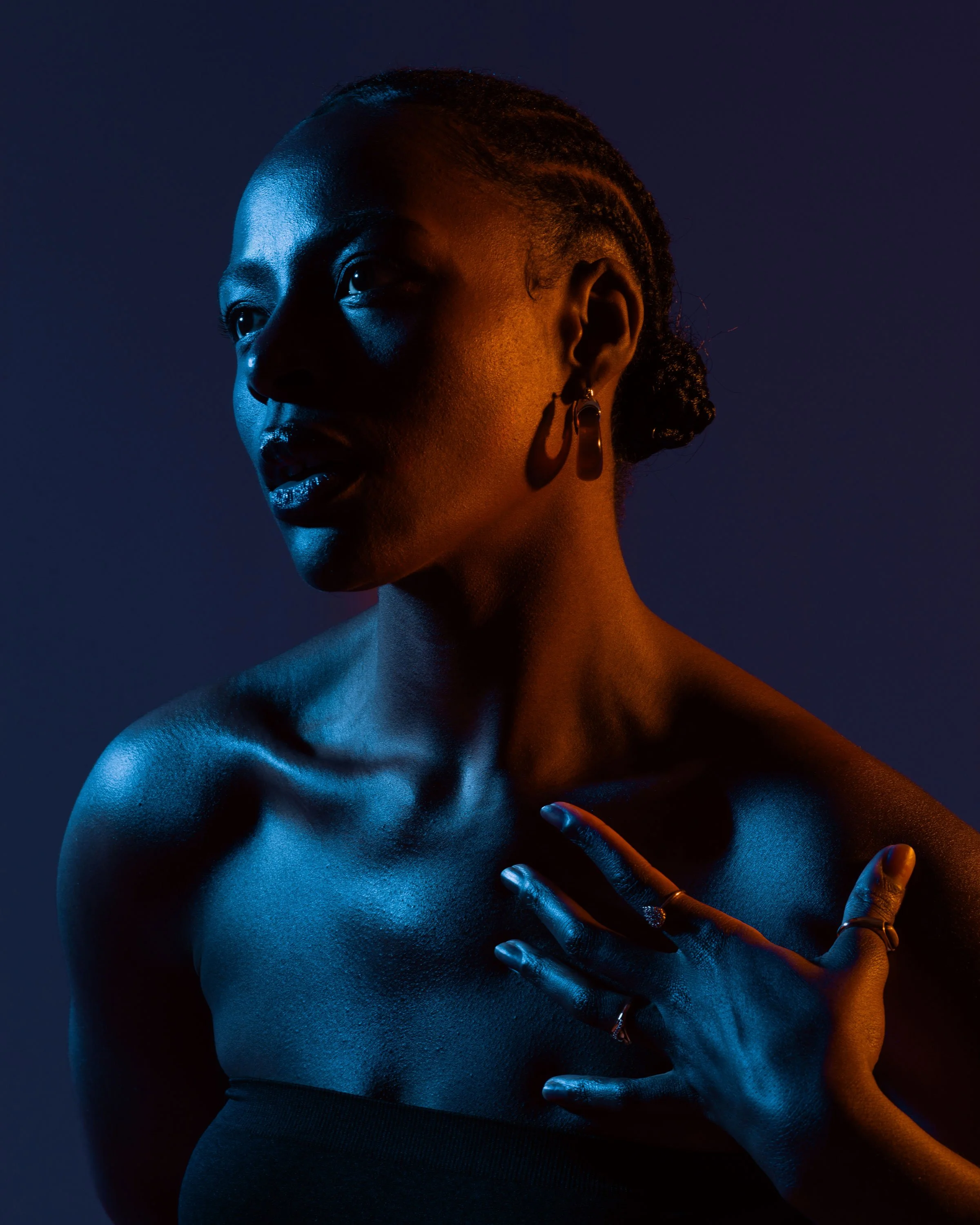 A portrait of a woman with short, braided hair, illuminated with blue and orange lighting, wearing earrings and rings, with her hand on her chest.