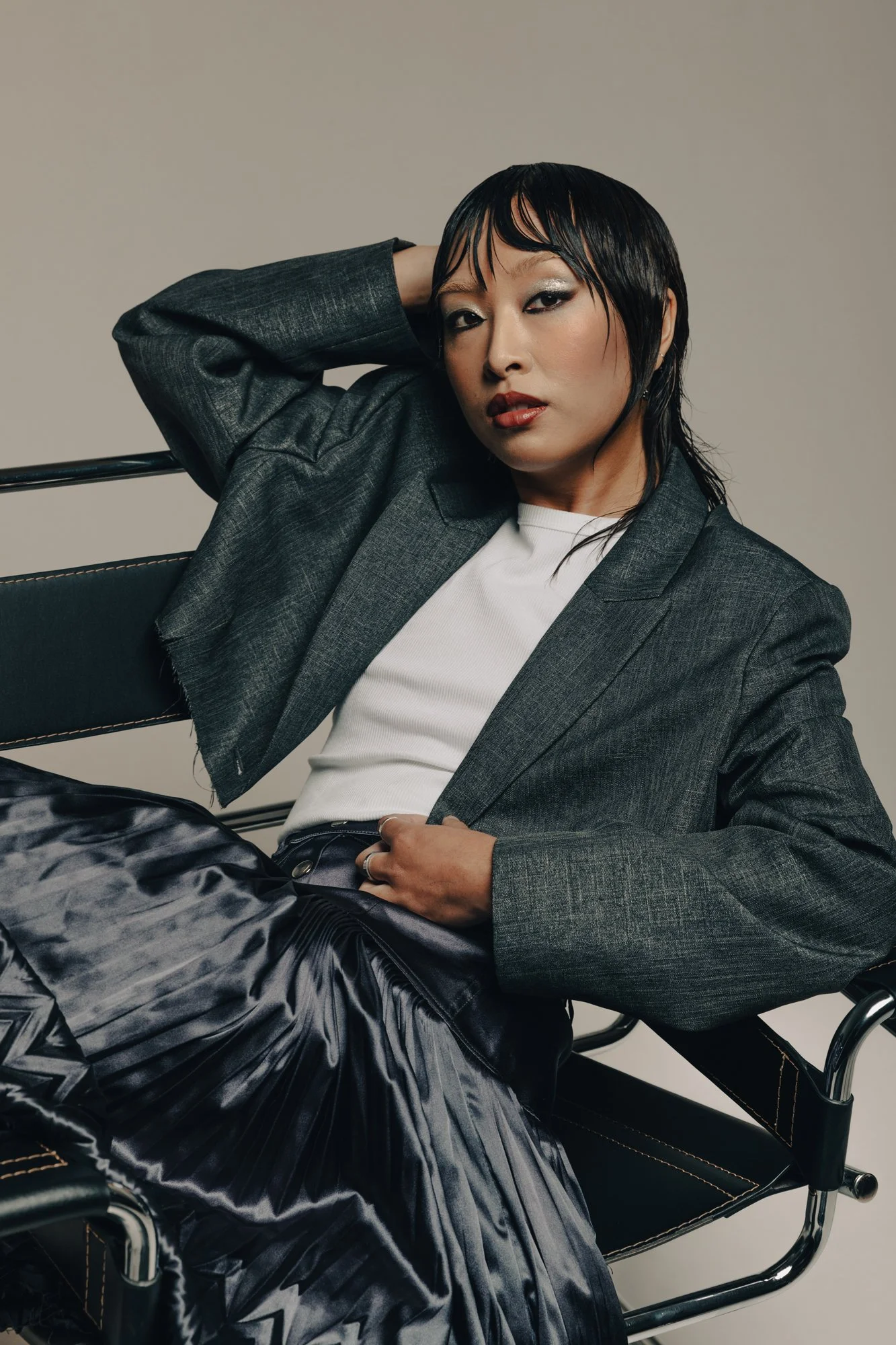 A woman with short black hair and makeup wearing a gray blazer, white top, and silky patterned skirt, sitting on a black chair against a plain background.