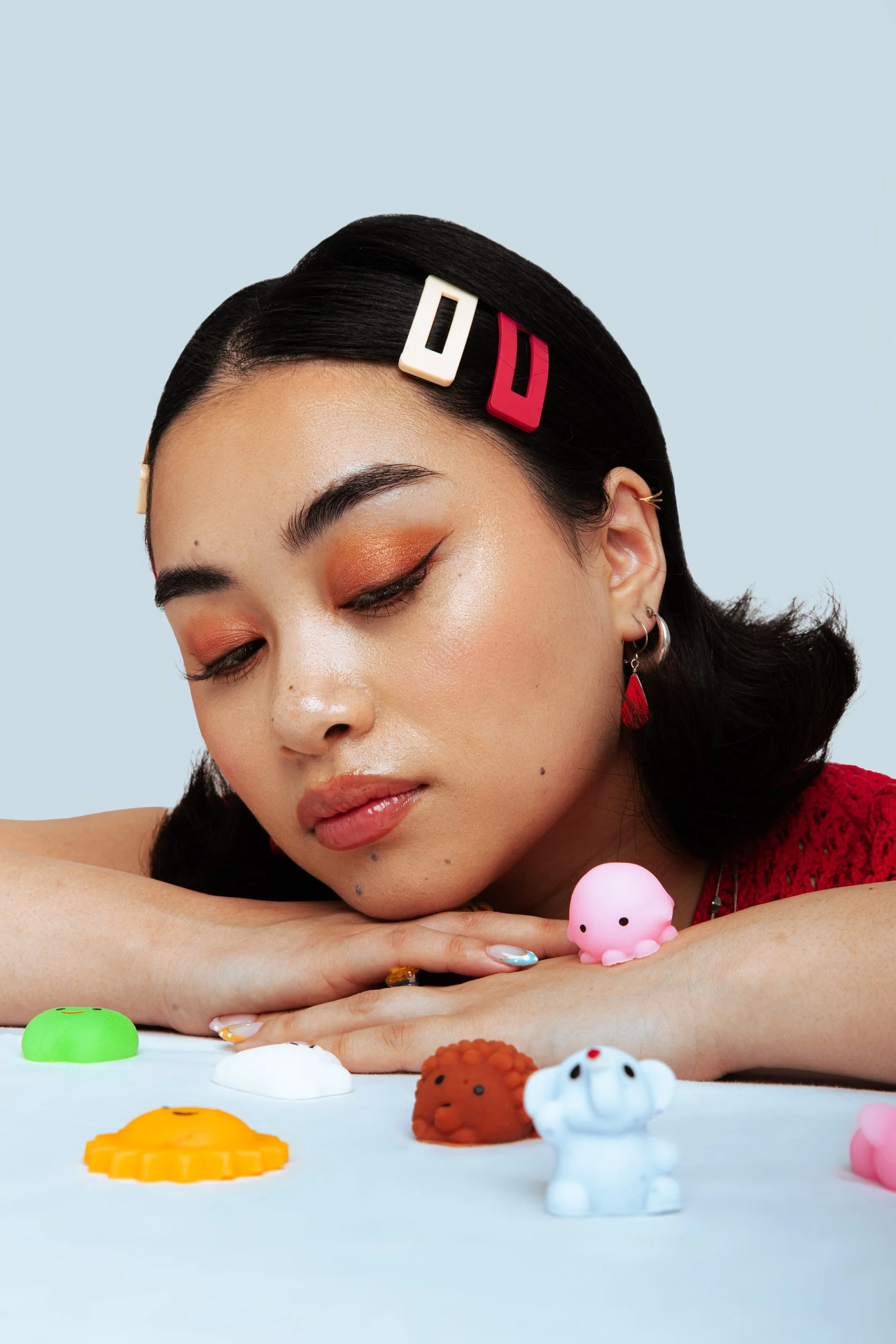 Young woman with black hair adorned with colorful hair clips, resting her face on her folded arms, surrounded by small cute figurines on a white surface, against a light blue background.