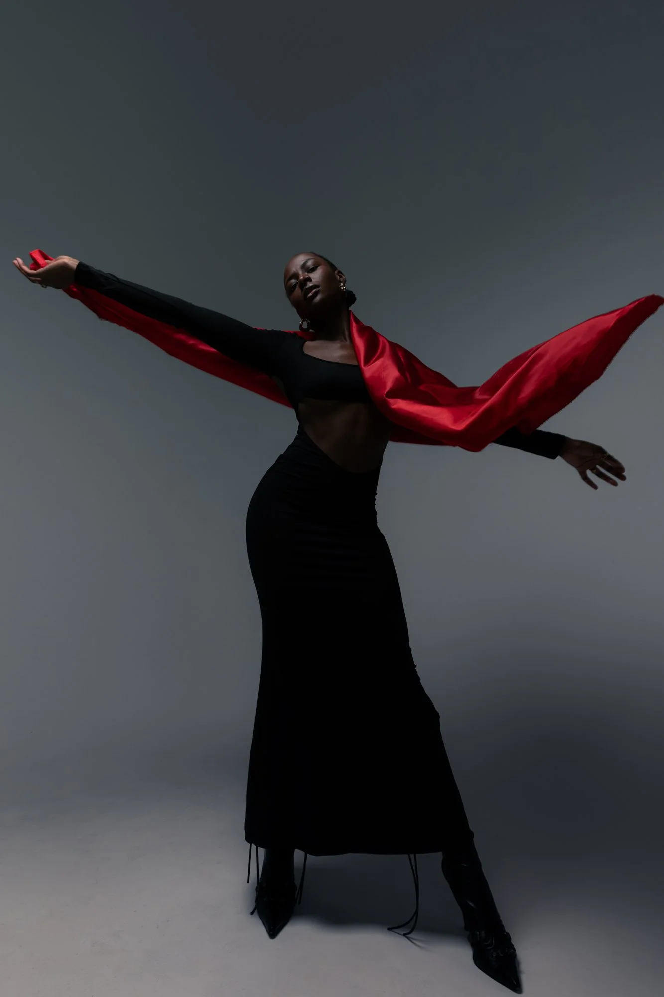 A woman in a black dress with sheer midsection, black high heels, and red satin cape stretches out her arms, posing dramatically against a gradient gray background.