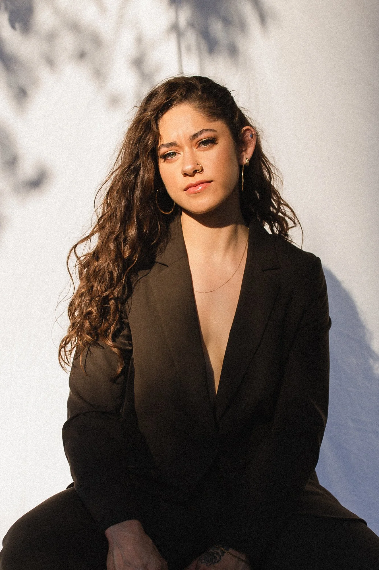 A young woman with long, curly brown hair and light skin, sitting outdoors against a plain white wall, wearing a black blazer and sports a nose piercing and hoop earrings.