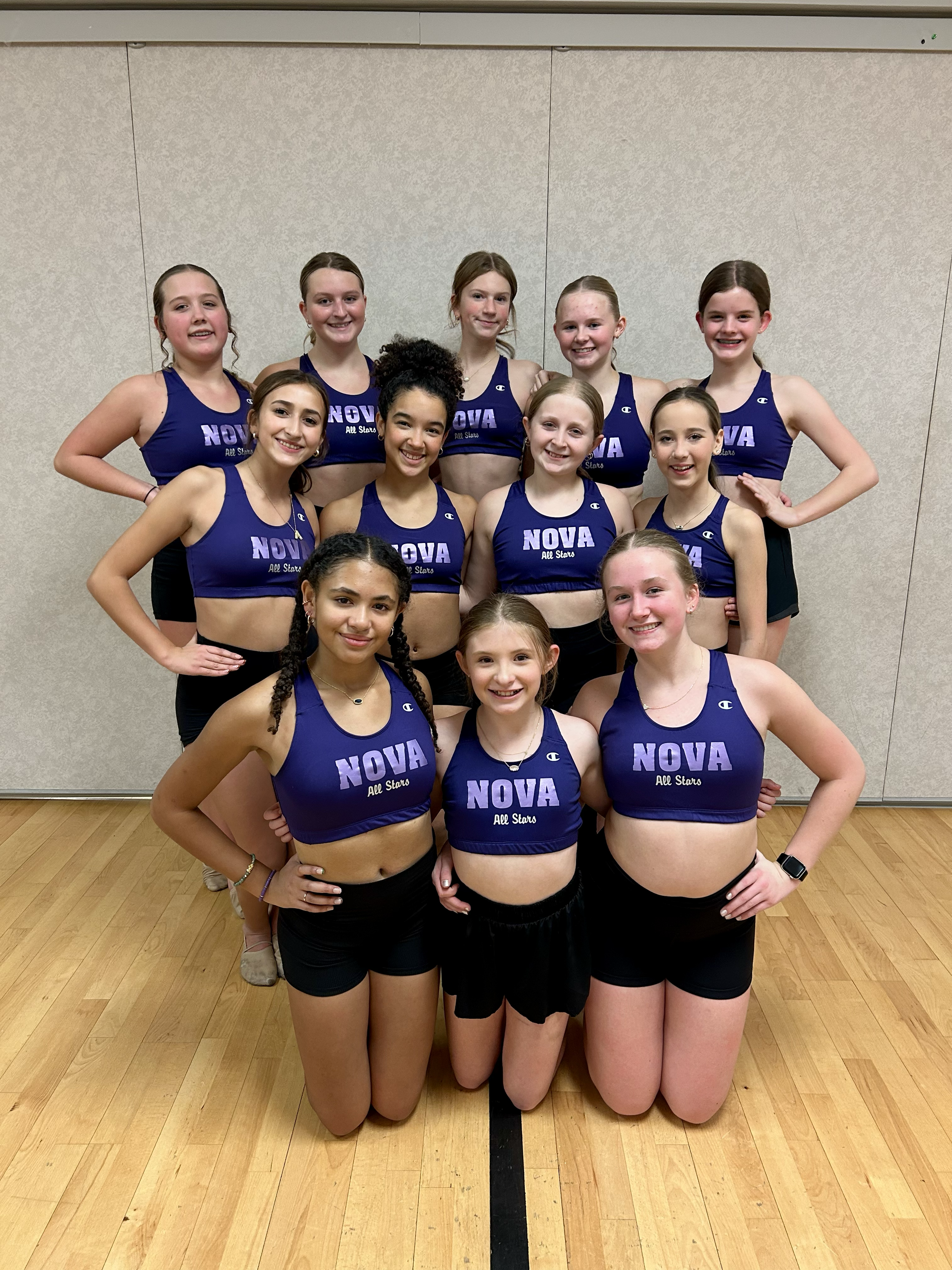 Group of young female athletes in blue sports bras with 'NOVA All Stars' logo, posing together indoors.