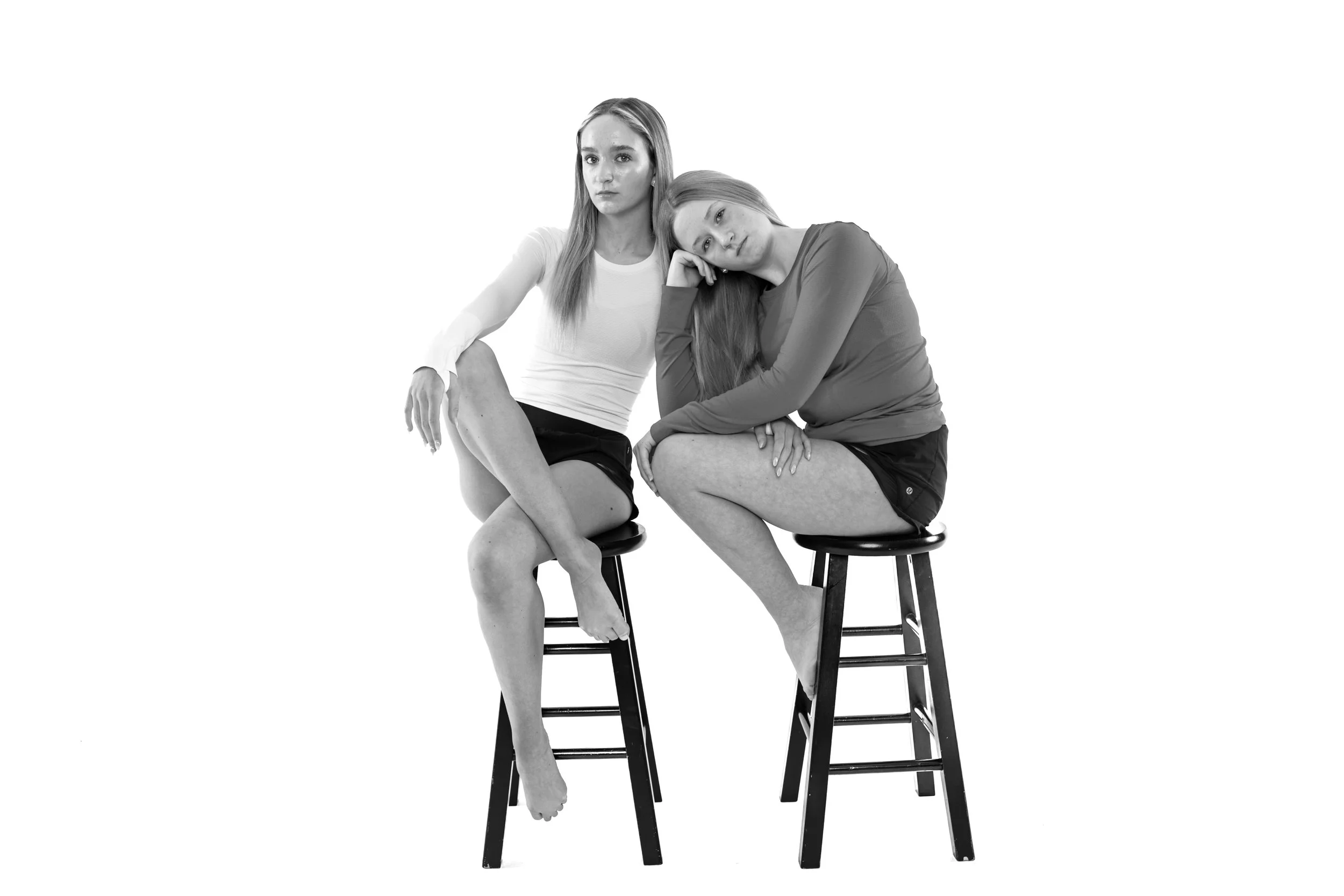 Two young women sitting on stools against a plain background, one resting her head on her hand and the other sitting with crossed legs, both wearing casual athletic clothing.