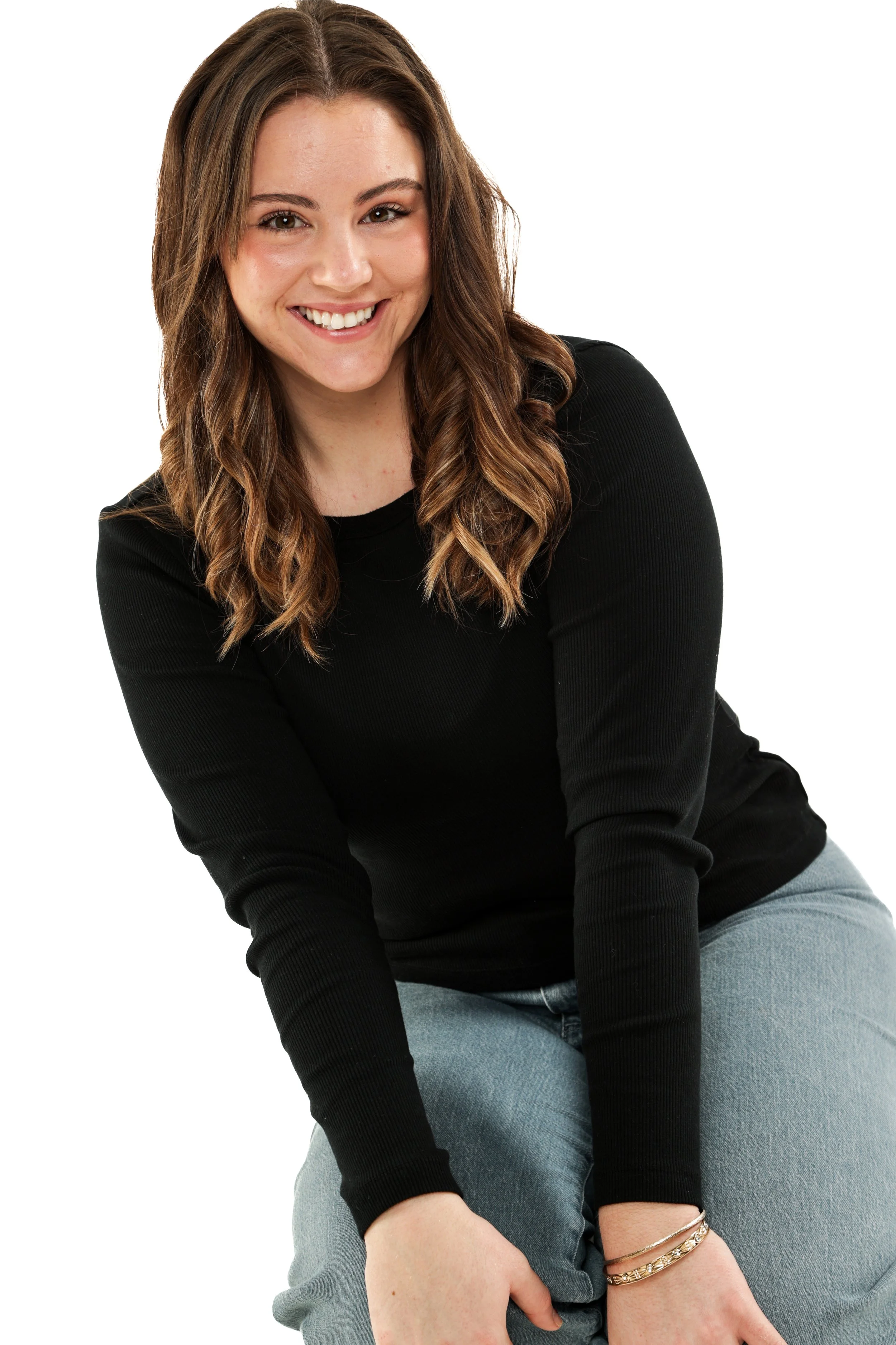A young woman with long brown hair wearing a black long-sleeve shirt and light blue jeans, smiling at the camera against a white background.