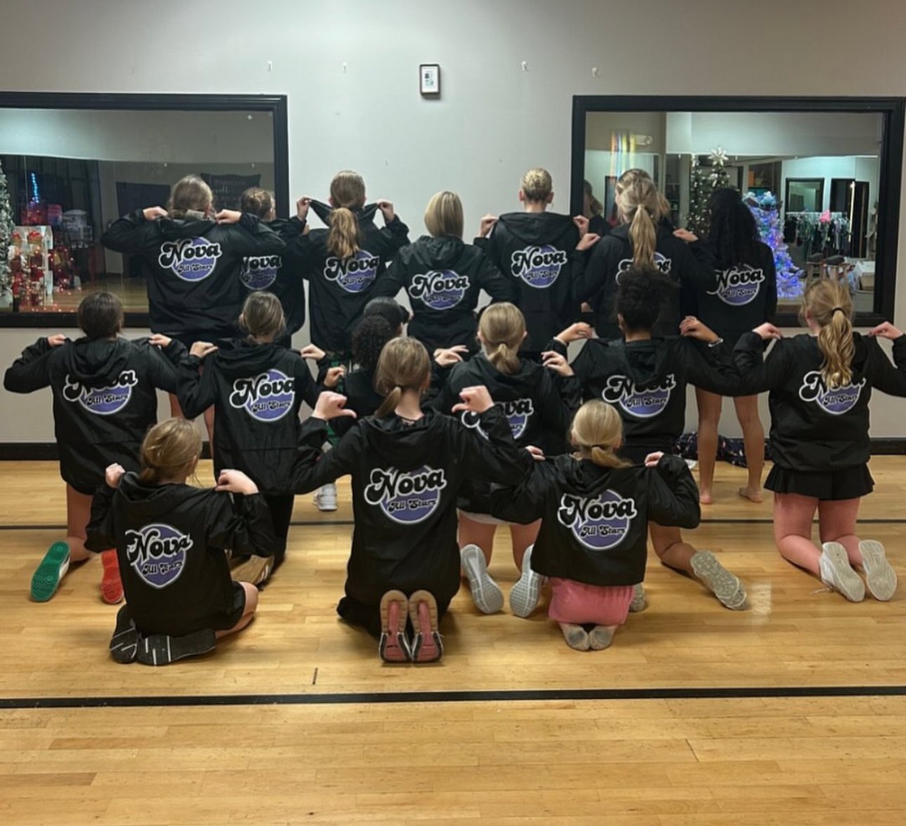 Group of young cheerleaders in black jackets with "Nova" logo, kneeling and standing, posing with arms flexed in gym or studio with wood floor, windows showing Christmas decorations and a decorated tree.