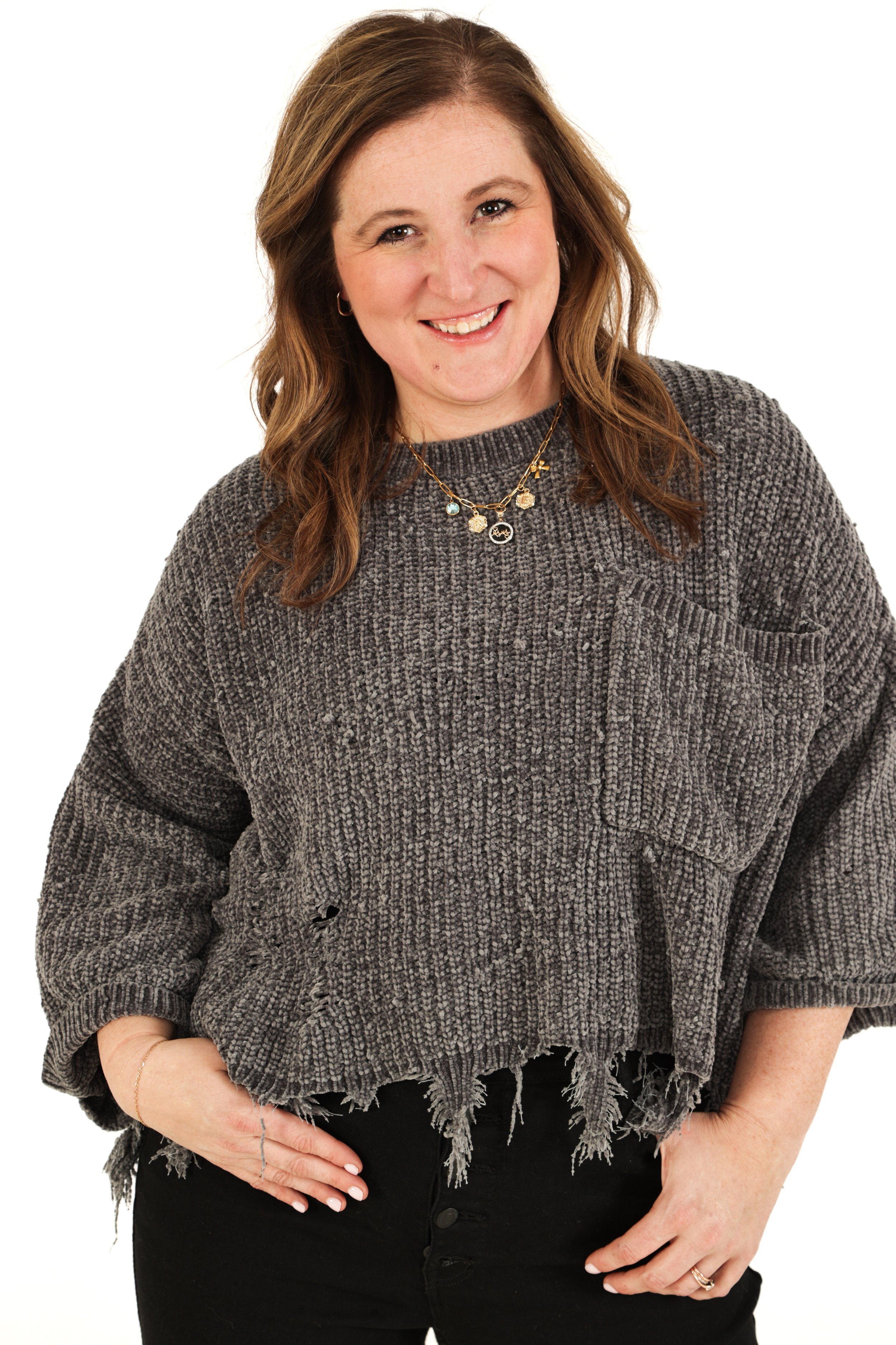A woman with shoulder-length brown hair, smiling, wearing a gray distressed sweater and black pants, accessorized with layered necklaces and a ring.