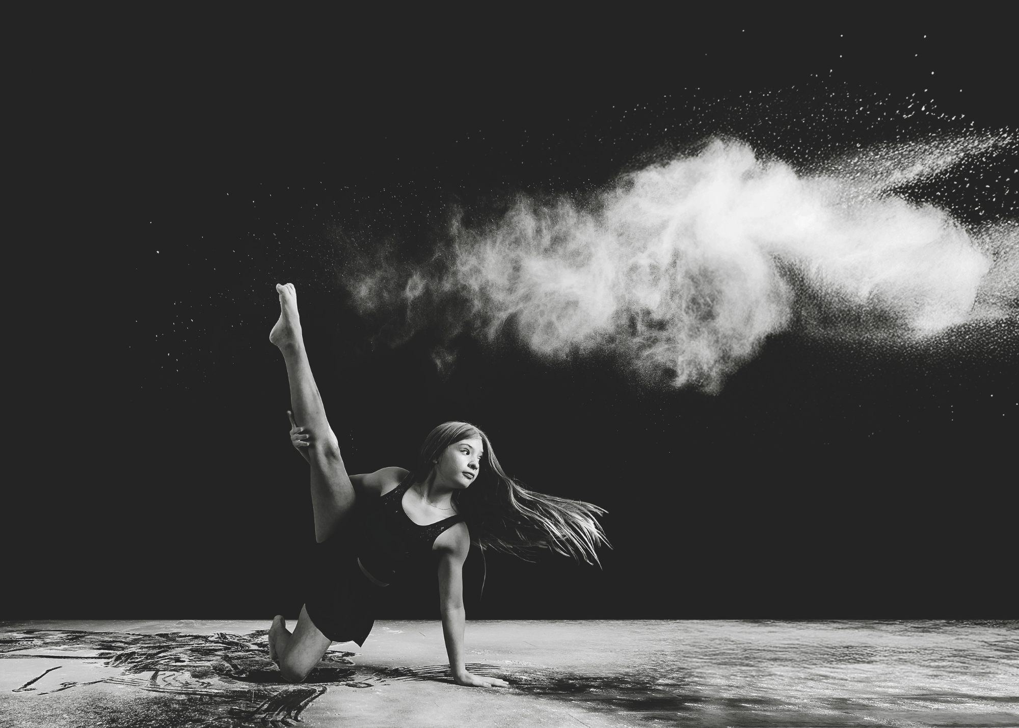 A woman in athletic clothing performing a one-legged yoga pose on the ground at night, with powder dispersing in the air.
