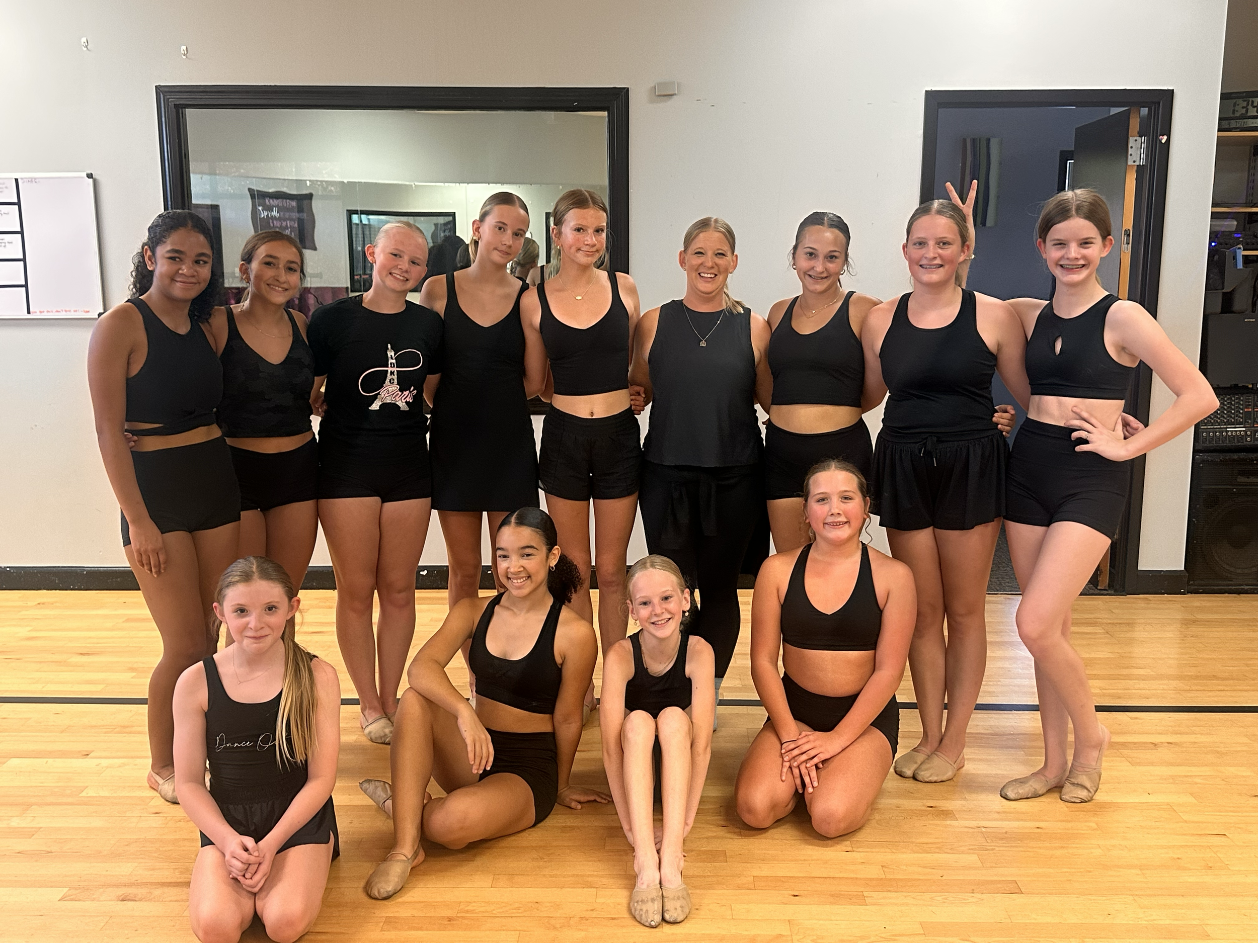 A group of young girls and their dance instructor posing for a photo in a dance studio. The girls are wearing black dance outfits, and the instructor is dressed in black as well. They are smiling and standing on a wooden floor with a mirror and a whi