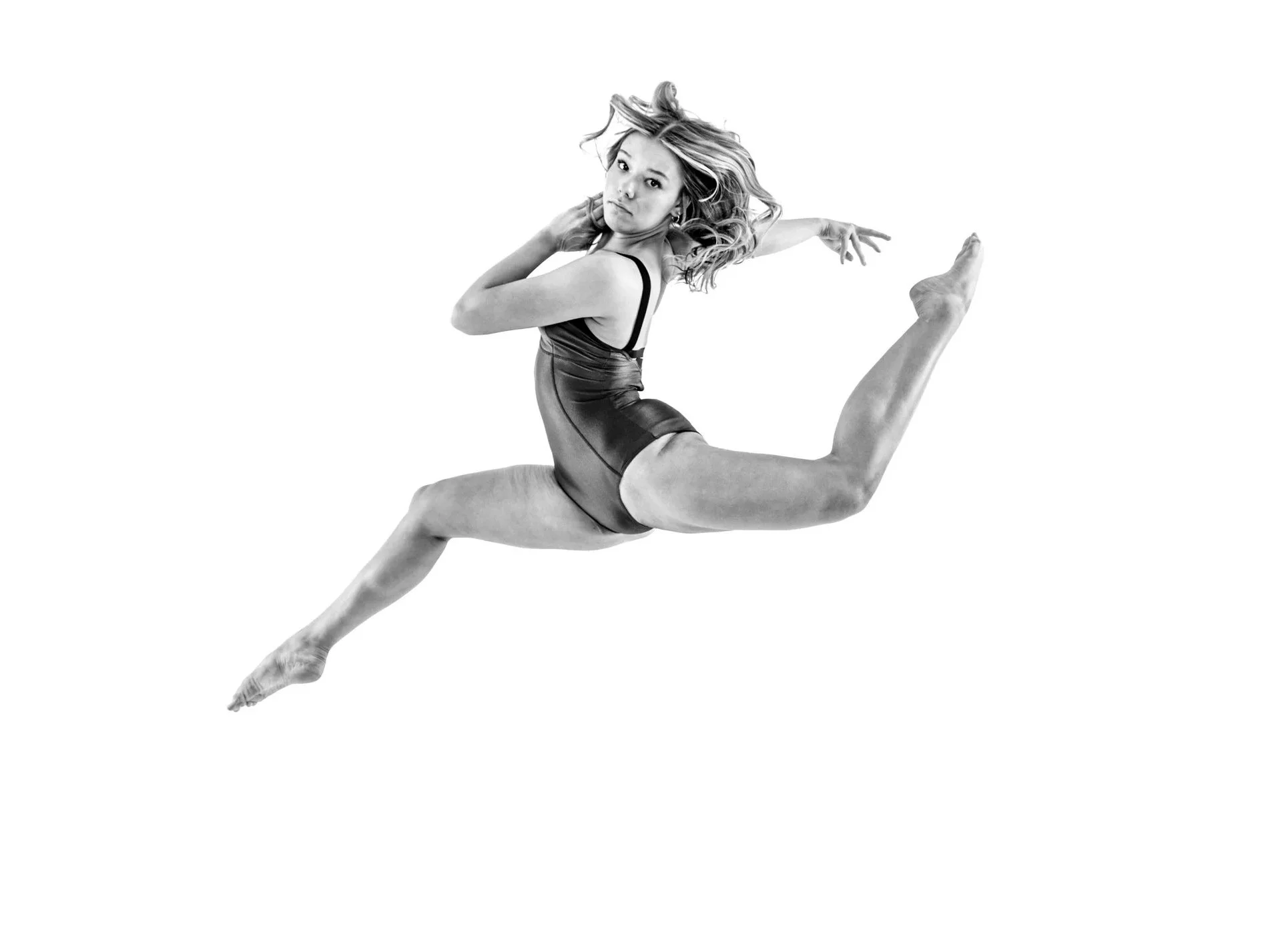 A woman in a dance pose wearing a black leotard, with her hair flowing, captured mid-air against a white background.