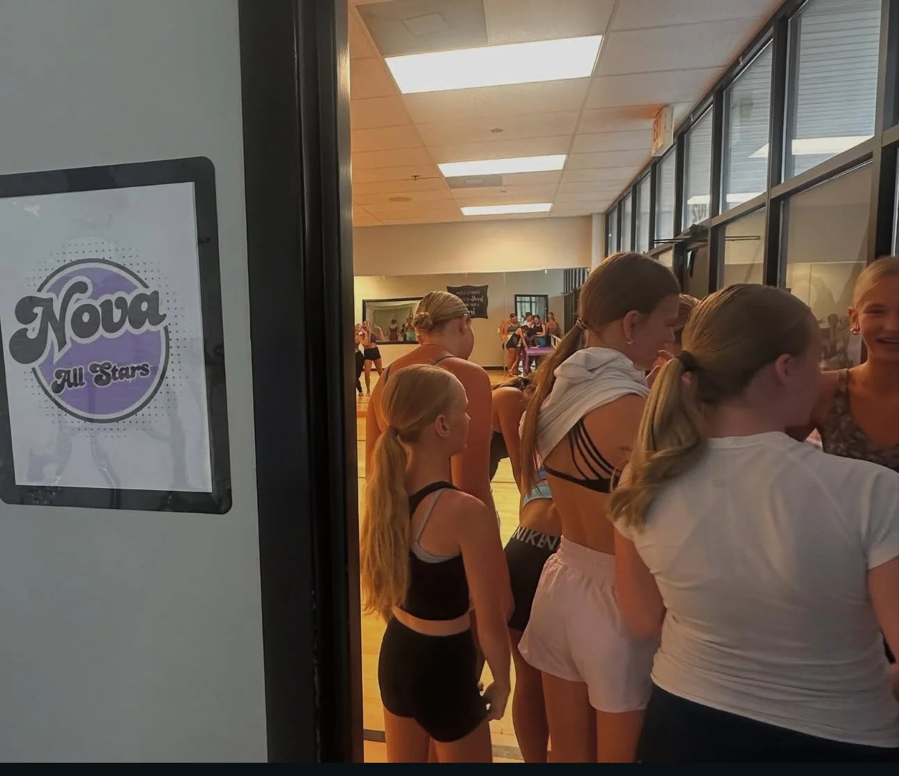 A group of young female gymnasts, dressed in athletic wear, waiting in line inside a dance studio or gym, with mirrors and other gymnasts visible in the background.