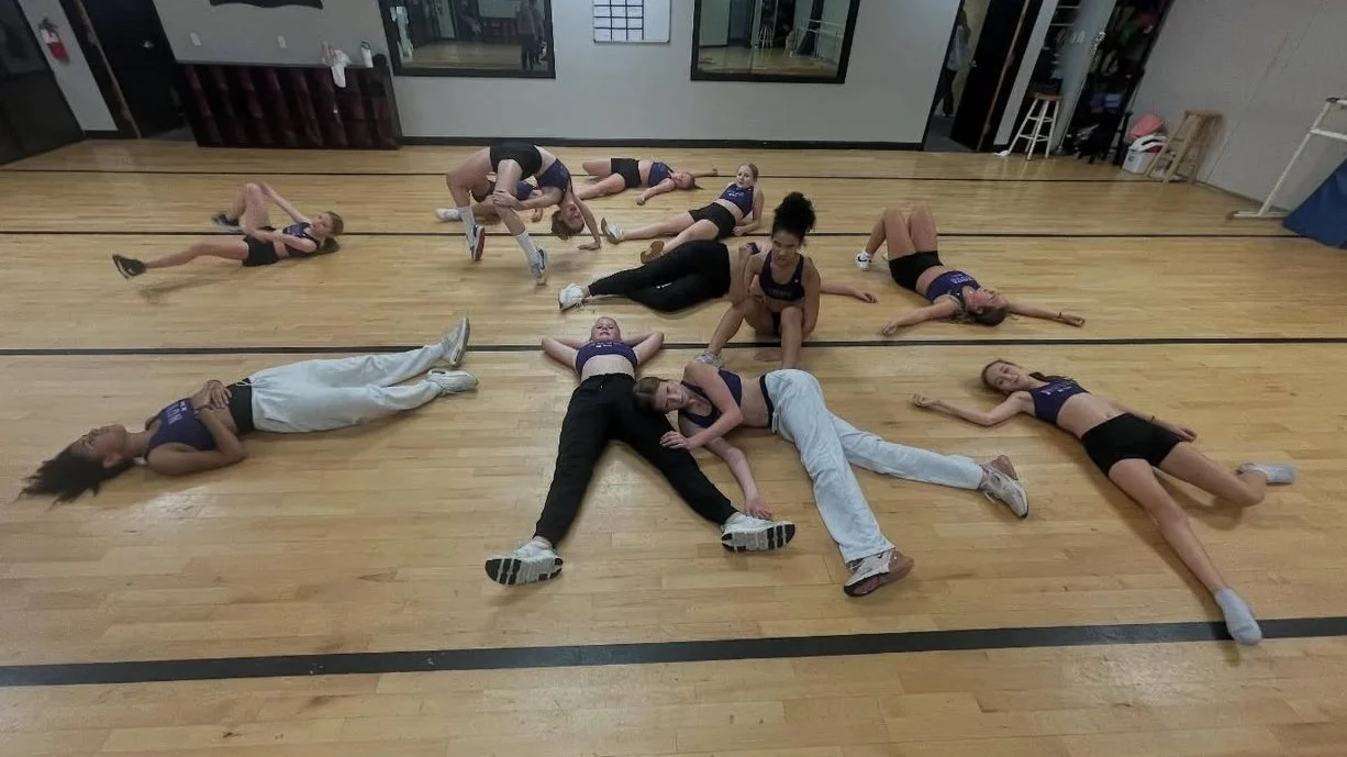 Group of young girls in athletic clothing lying and sitting on a dance studio floor, some posing and some resting.