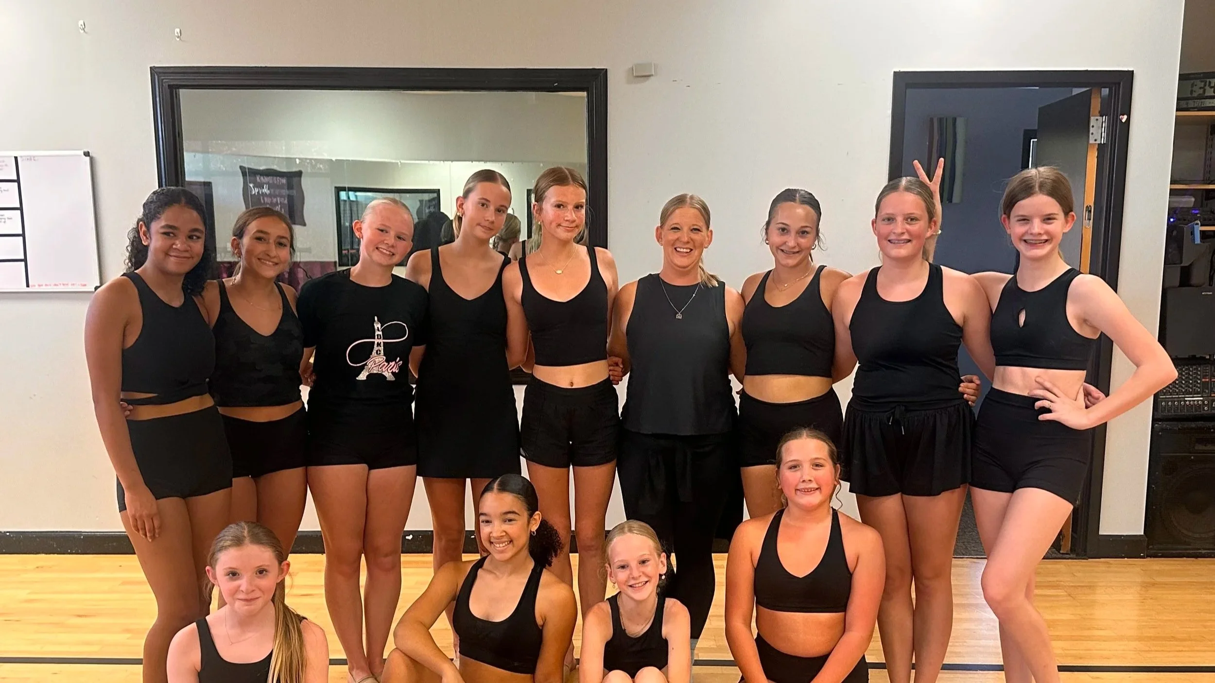 Group of young girls and their dance instructor pose for a photo in a dance studio, all wearing black dance attire. The instructor stands in the center, smiling, with girls standing and sitting around her.