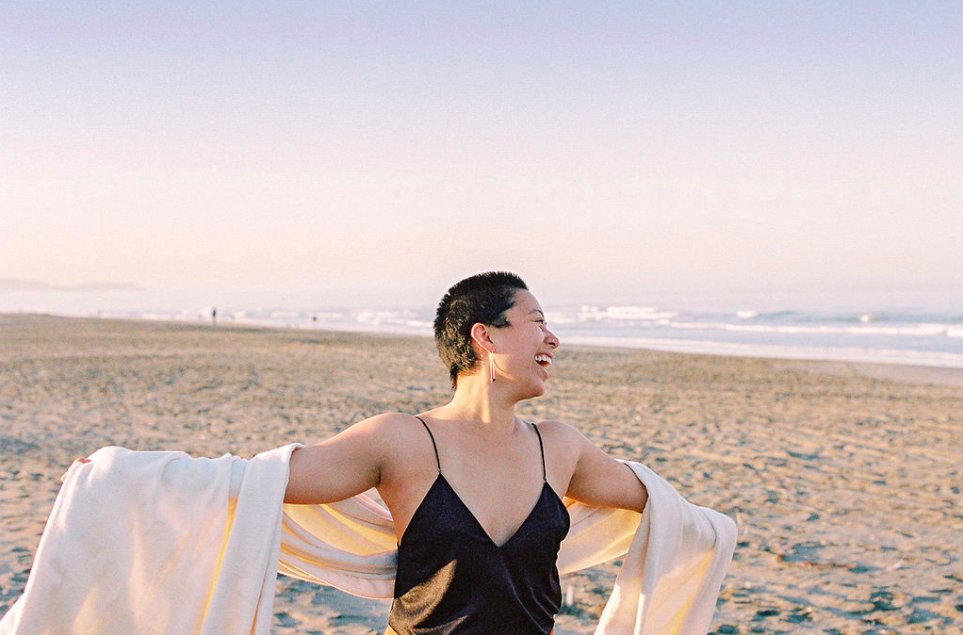 Trauma-informed somatic practitioner with short hair and earrings smiling on a sandy beach at sunset, wearing a black dress and holding a light-colored jacket.