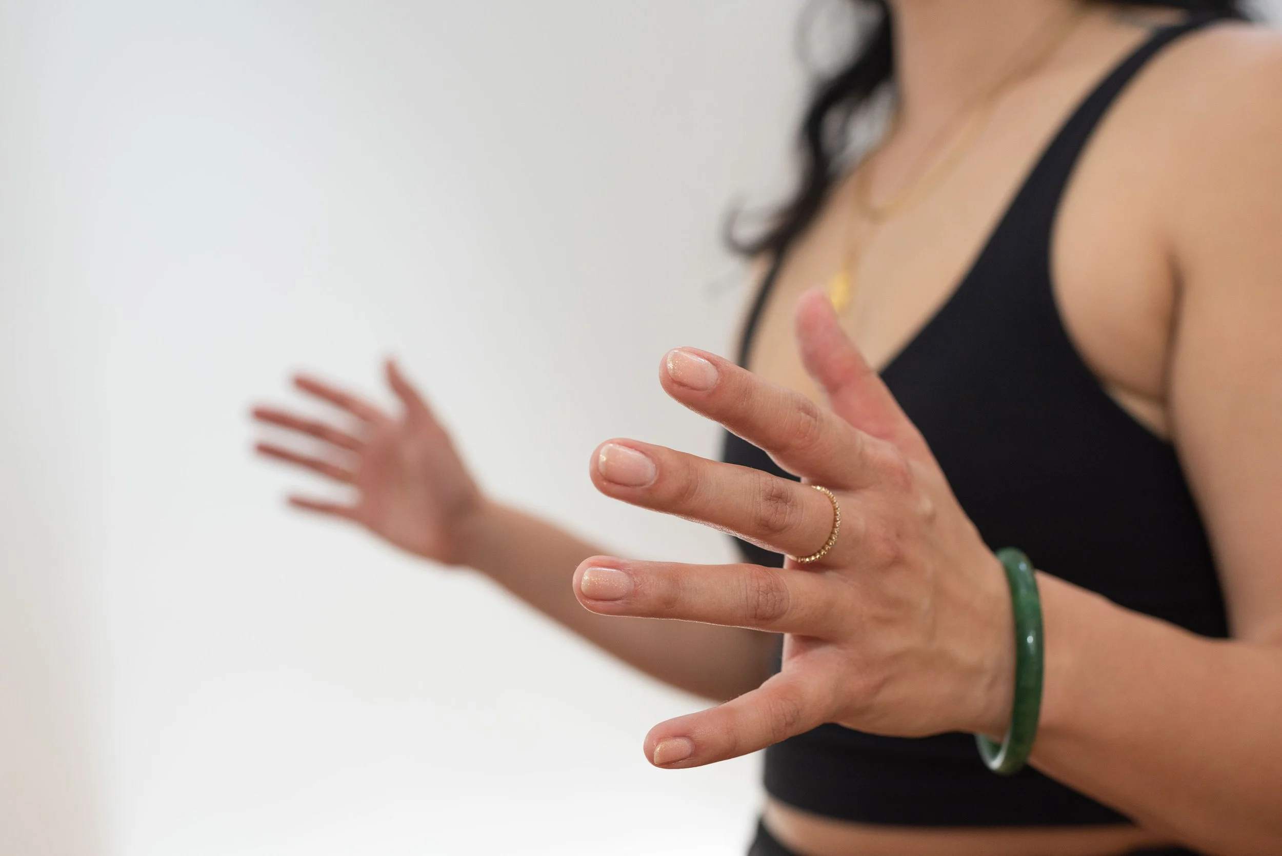 A woman with dark hair wearing a black tank top, jewelry including a green bracelet, a gold ring, and a necklace, with her hands raised and fingers slightly spread apart.