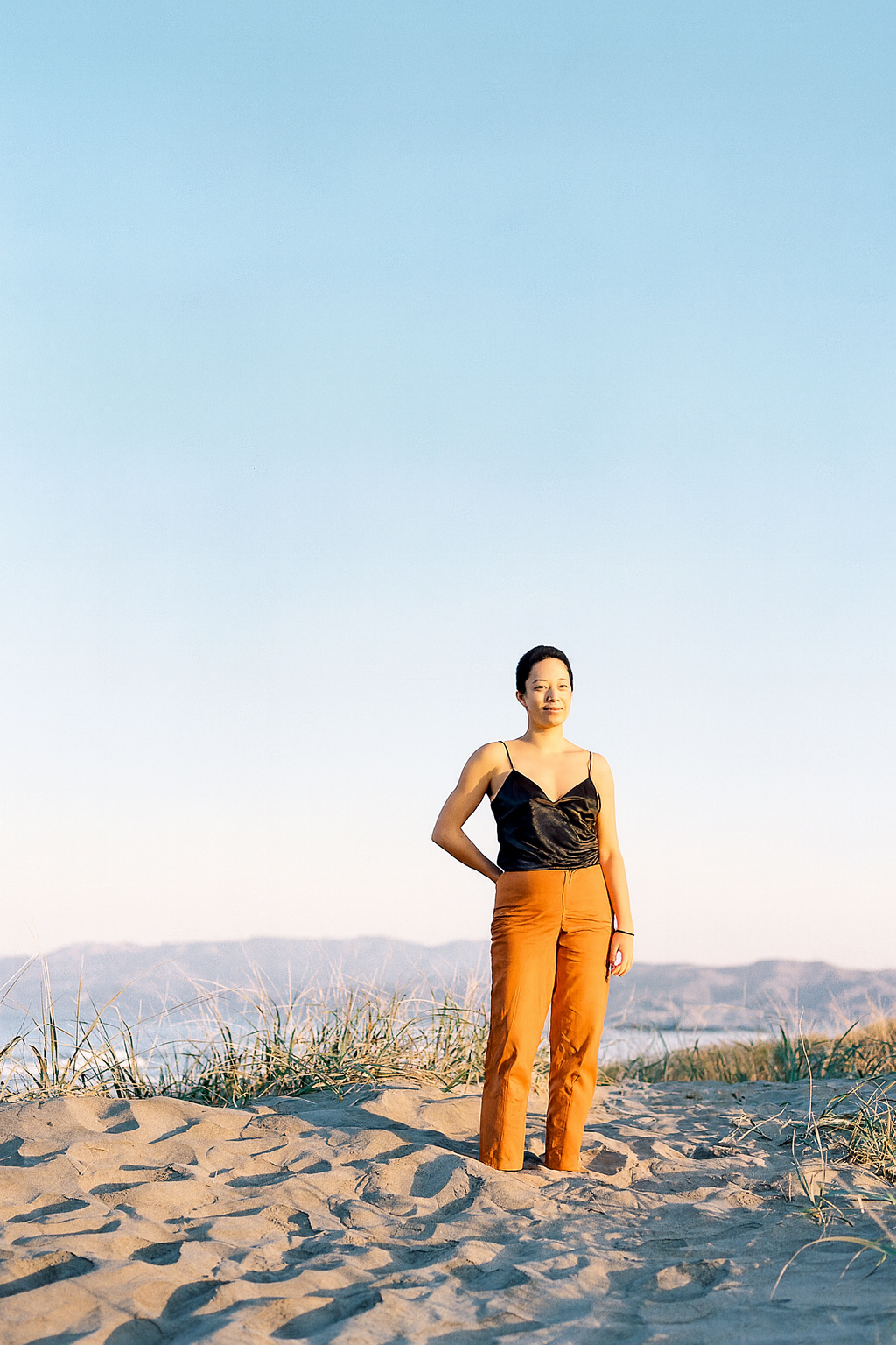 A woman standing on sandy beach with grassy dunes and distant mountains under a clear blue sky, embodying calm somatic healing practice.