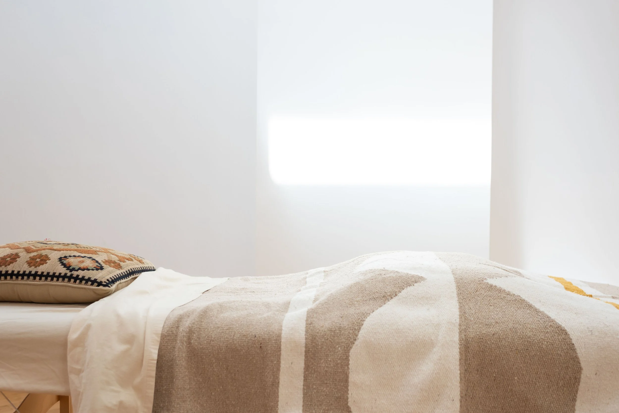 A neatly made bed with a decorative pillow and a patterned beige and white blanket, positioned against a plain white wall with a small window emitting bright light.