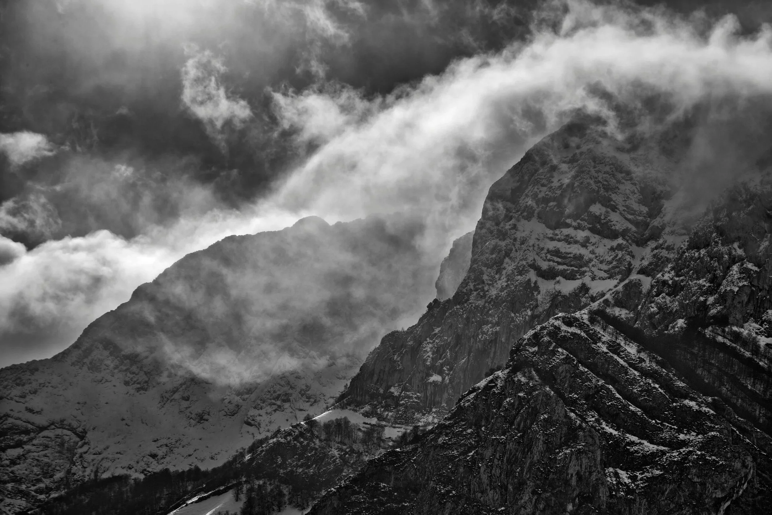 Snow-covered mountain peaks with clouds swirling around them in black and white, illustrating the ruggedness of life's terrain that is explored within a trauma informed, liberatory framework session.