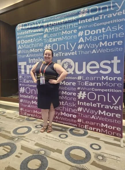 Woman standing in front of a blue backdrop with hashtags and text related to a tech or travel event. She is wearing a black dress and sandals, and has a conference badge hanging around her neck.