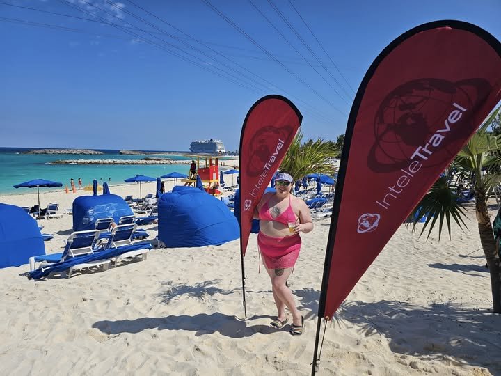 A woman in a pink swimsuit and cover-up standing on a beach next to two red flags with white text and logos, with blue lounge chairs, umbrellas, a lifeguard chair, and a cruise ship in the background on a sunny day.