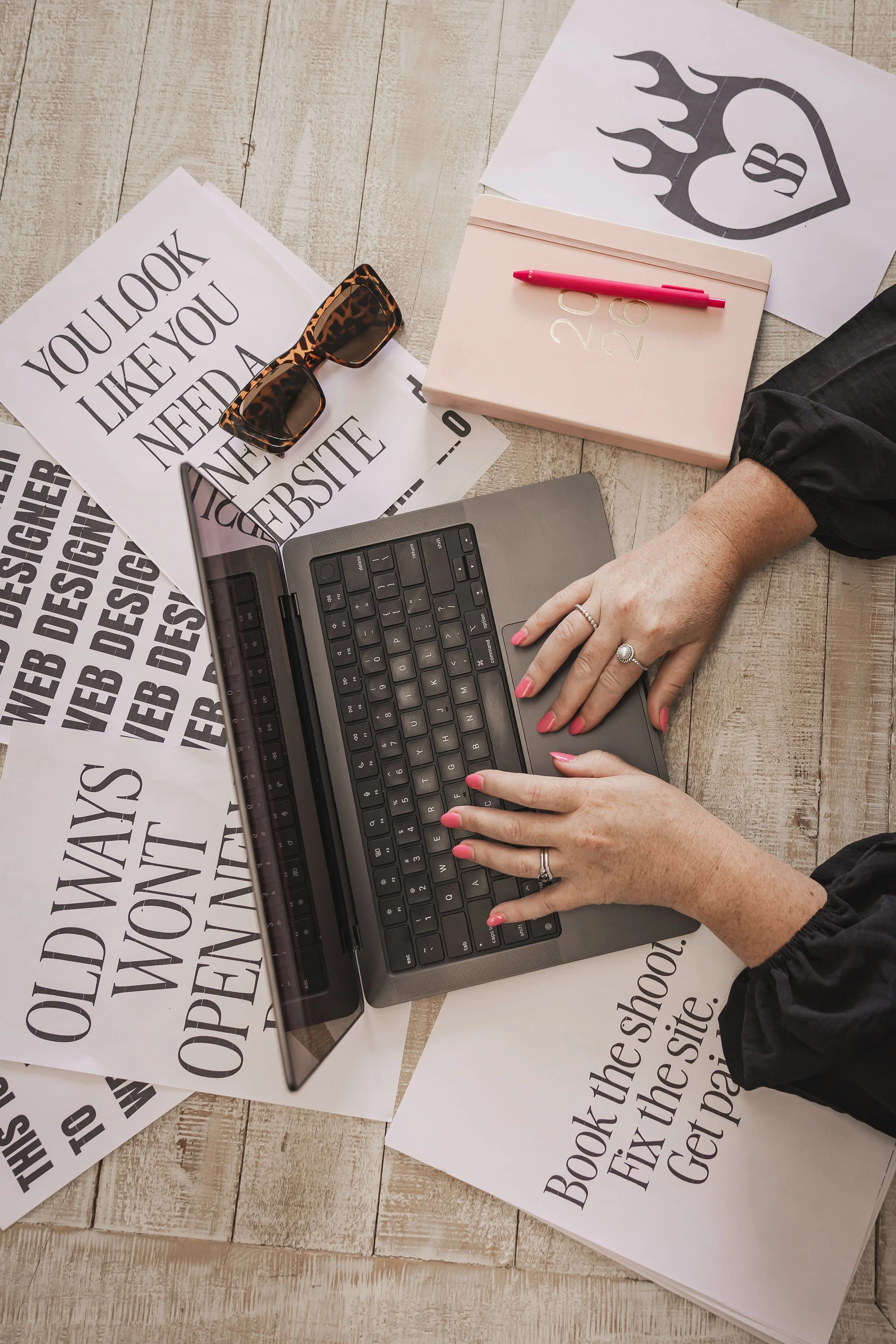 A person working on a laptop surrounded by printed papers with bold text, a pink planner with a pink pen on top, a pair of sunglasses, and eye-catching artwork with a heart and flames.