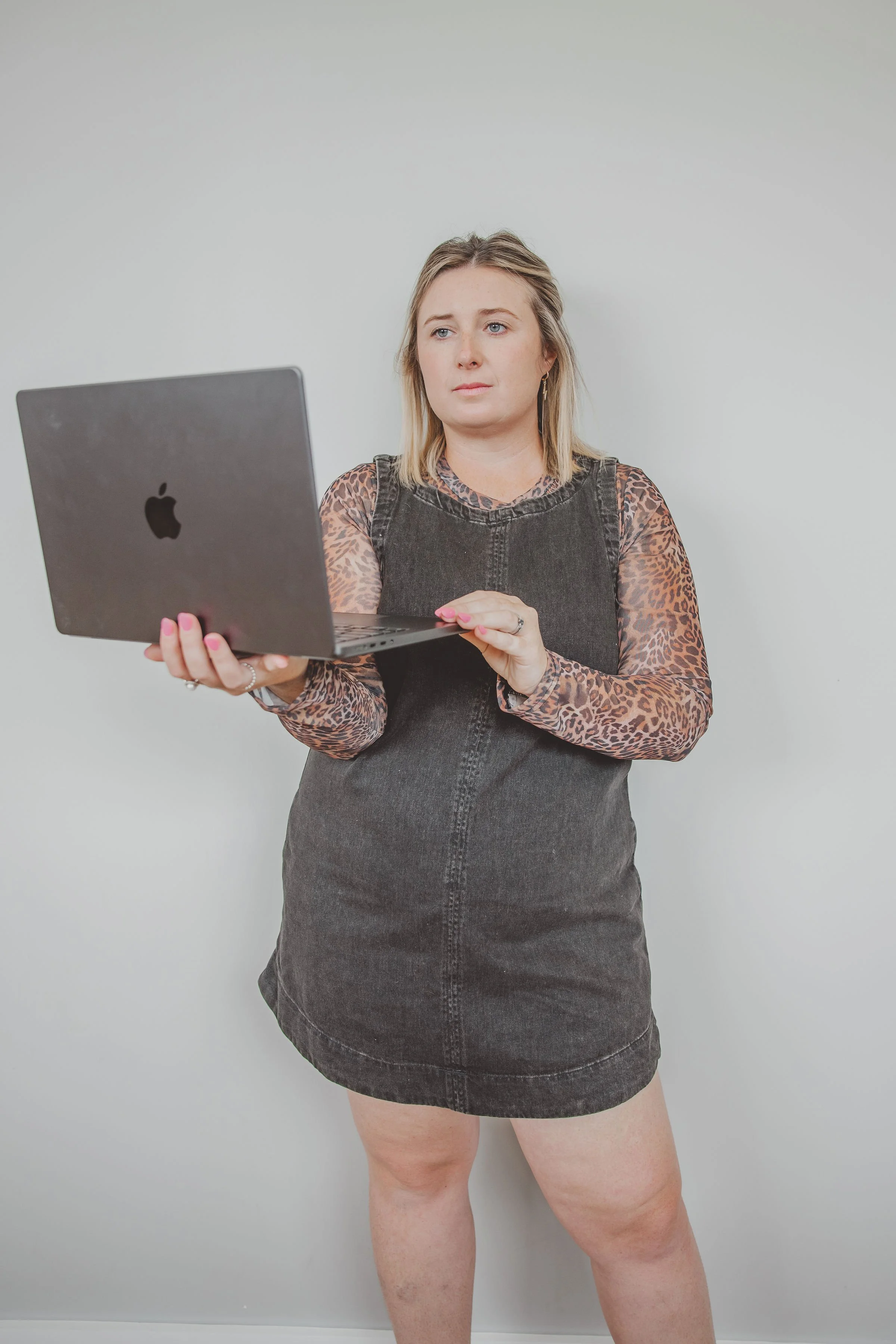 A woman with blonde hair holding a silver MacBook laptop, wearing a black denim dress with leopard print long sleeves, standing against a plain white wall.