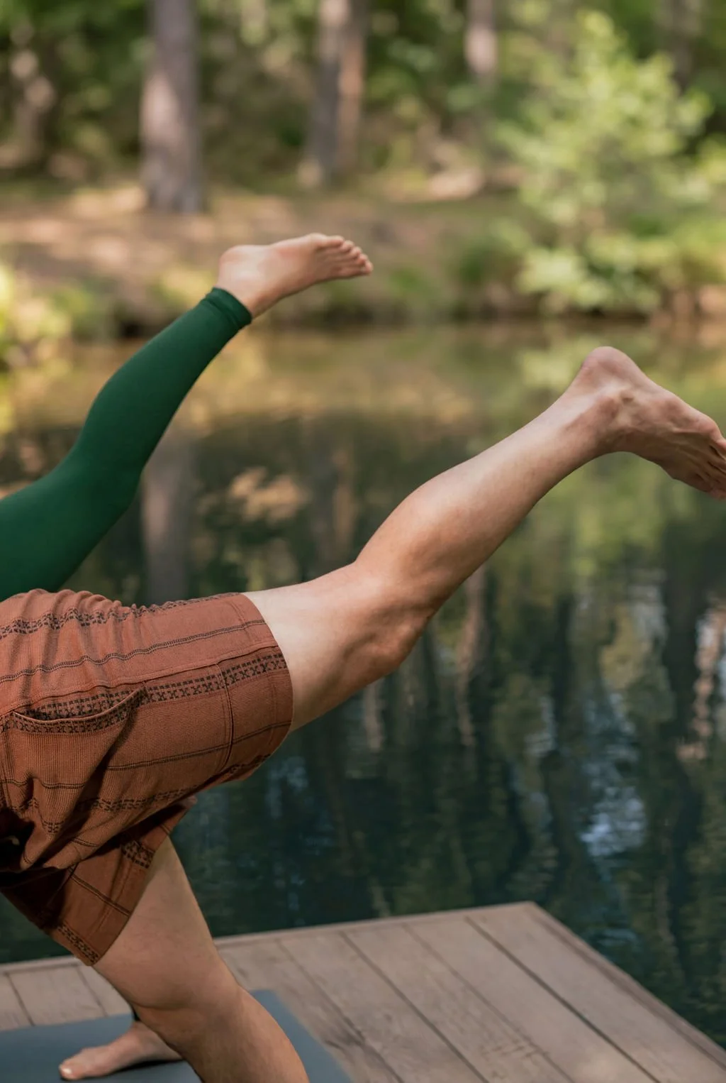 Person practicing yoga in a one-legged pose on a dock near a pond in a forested area.