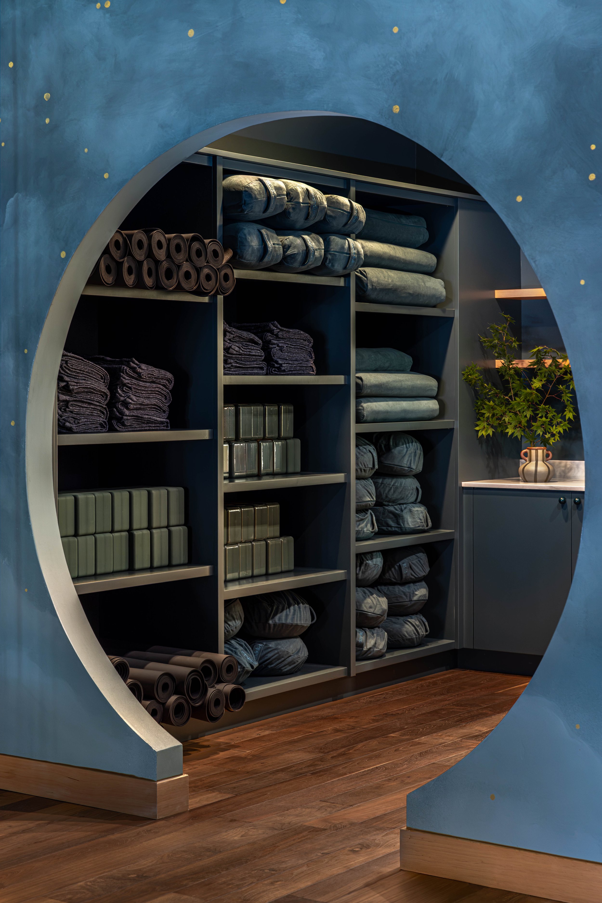 Shelves of folded towels, yoga mats, and blankets in a fitness or yoga studio with blue walls and wooden flooring.