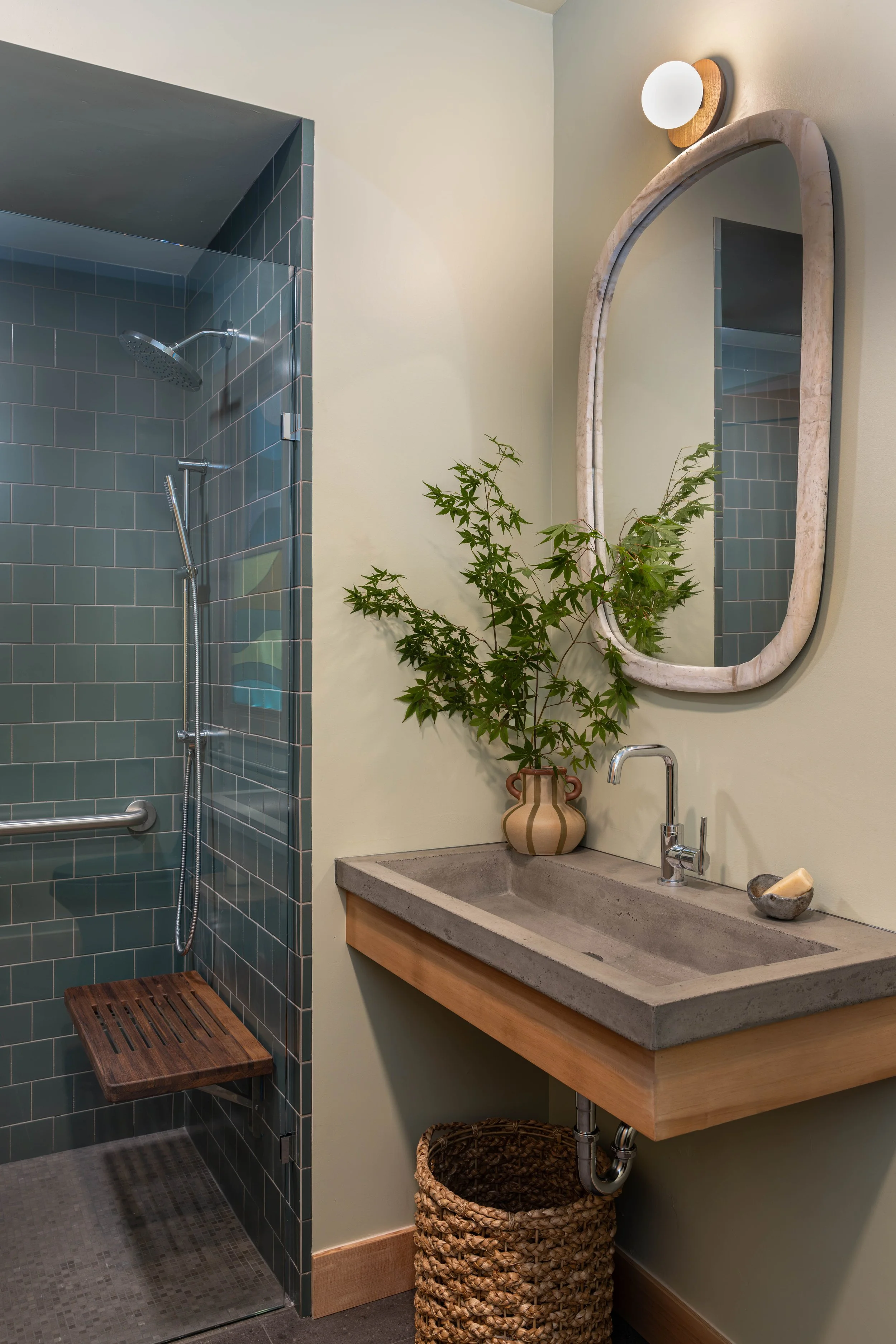 Modern bathroom with a walk-in shower featuring teal tiles and a wooden bench. A concrete sink with a chrome faucet, a large mirror with a curved frame, a plant in a striped vase, and a wicker basket in the corner.